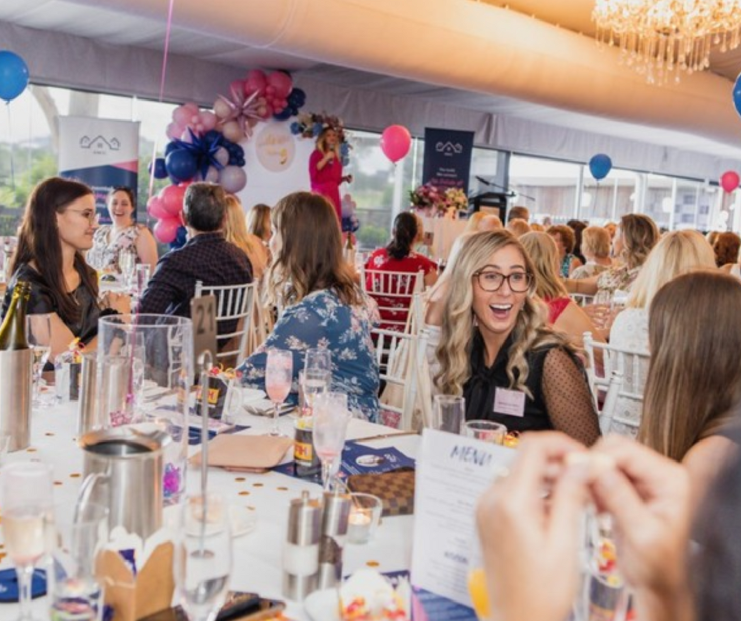 People seated at decorated tables with glasses, plates, and menus, while a speaker addresses the audience from a podium under a chandelier. Blue and pink balloons adorn the room, indicating a formal event. Prop hire, Prop hiring, Prop hire, event stylists Brisbane, brisbane event styling, brisbane event styling, event stylist Brisbane, Brisbane Wedding, Gold Coast Wedding, Floral Styling, Lush & Lavish Events, Brisbane Corporate Styling, Gold Coast Corporate Styling, Brisbane Brand Activation