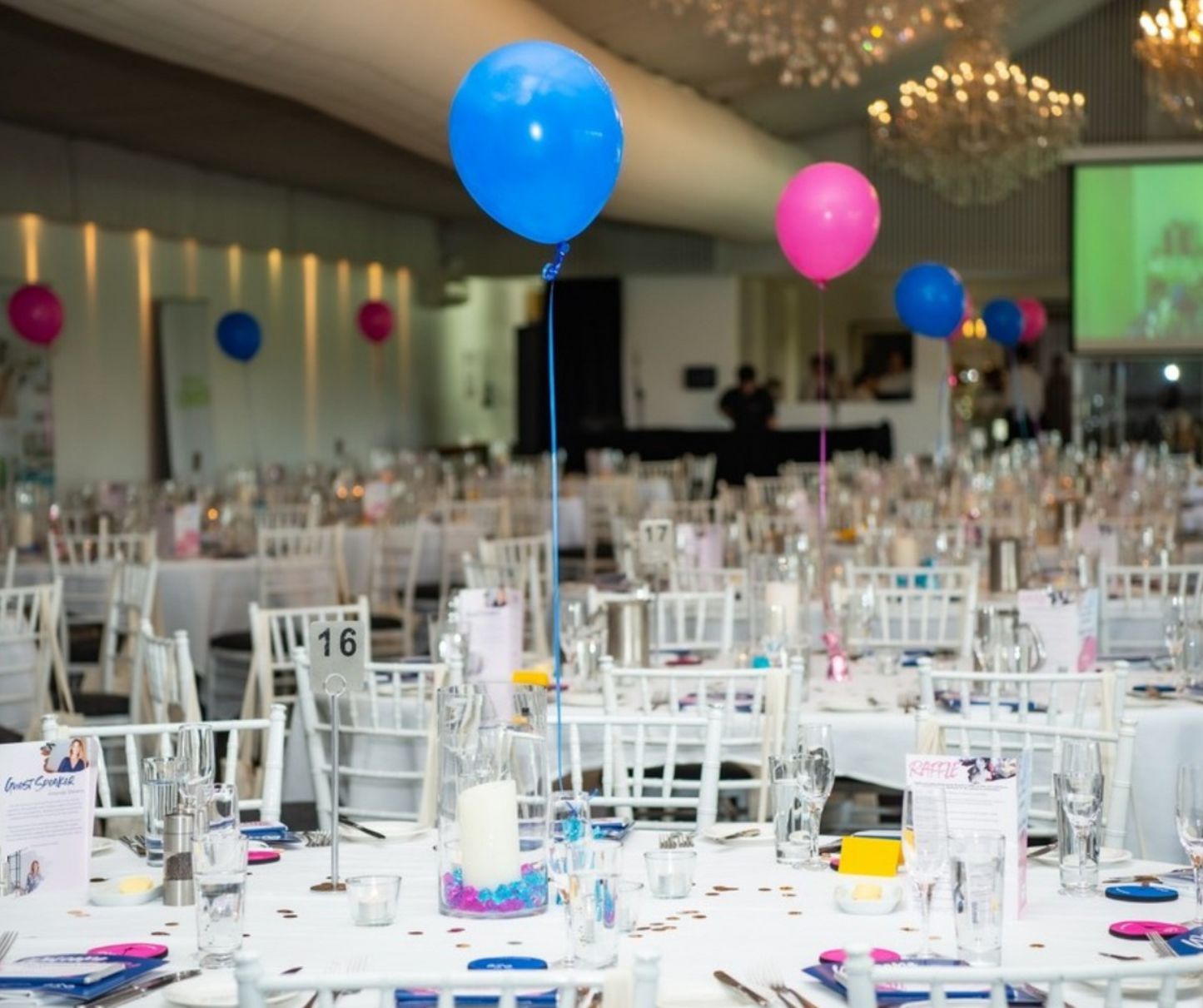An indoor event features white-clothed tables, blue and pink balloon centerpieces, chandeliers, and a presentation screen. This event was styled by Lush & Lavish Events, specializing in Brisbane and Gold Coast events, weddings, and corporate functions. Prop hire, Prop hiring, Prop hire, event stylists Brisbane, brisbane event styling, brisbane event styling, event stylist Brisbane, Brisbane Wedding, Gold Coast Wedding, Floral Styling, Lush & Lavish Events, Brisbane Corporate Styling, Gold Coast Corporate St