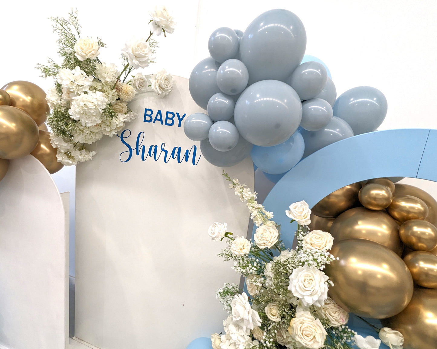 The image shows a decorative setup for a baby celebration event. The setup includes a white backdrop with the text "BABY Sharan" written on it. Surrounding the backdrop are clusters of balloons in shades of blue and gold, along with arrangements of white flowers. The decorations are elegant and festive, indicating a special occasion, likely a baby shower or a welcome party for a newborn named Sharan. Prop hire, Prop hiring, Prop hire, event stylists Brisbane, brisbane event styling, brisbane event styling