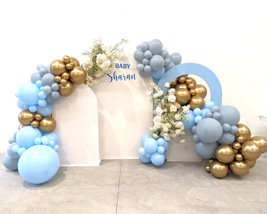 A decorative arch made of blue and gold balloons is set up against a white background. The arch is adorned with white flowers and has a sign that reads "BABY SHOWER" in blue letters. The balloons vary in size, creating a visually appealing arrangement. Prop hire, Prop hiring, Prop hire, event stylists Brisbane, brisbane event styling, brisbane event styling, event stylist Brisbane, Brisbane Wedding, Gold Coast Wedding, Floral Styling, Lush & Lavish Events