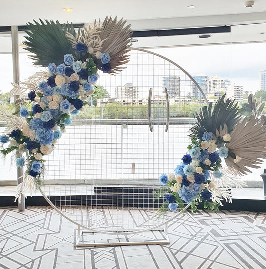 ircular floral arrangement indoors against a glass wall with a river and cityscape view. Features blue and white flowers, dried palm leaves, on a metal hoop stand, on a geometric patterned carpet. Prop hire, Prop hiring, Prop hire, event stylists Brisbane, brisbane event styling, brisbane event styling, event stylist Brisbane, Brisbane Wedding, Gold Coast Wedding, Floral Styling, Lush & Lavish Events