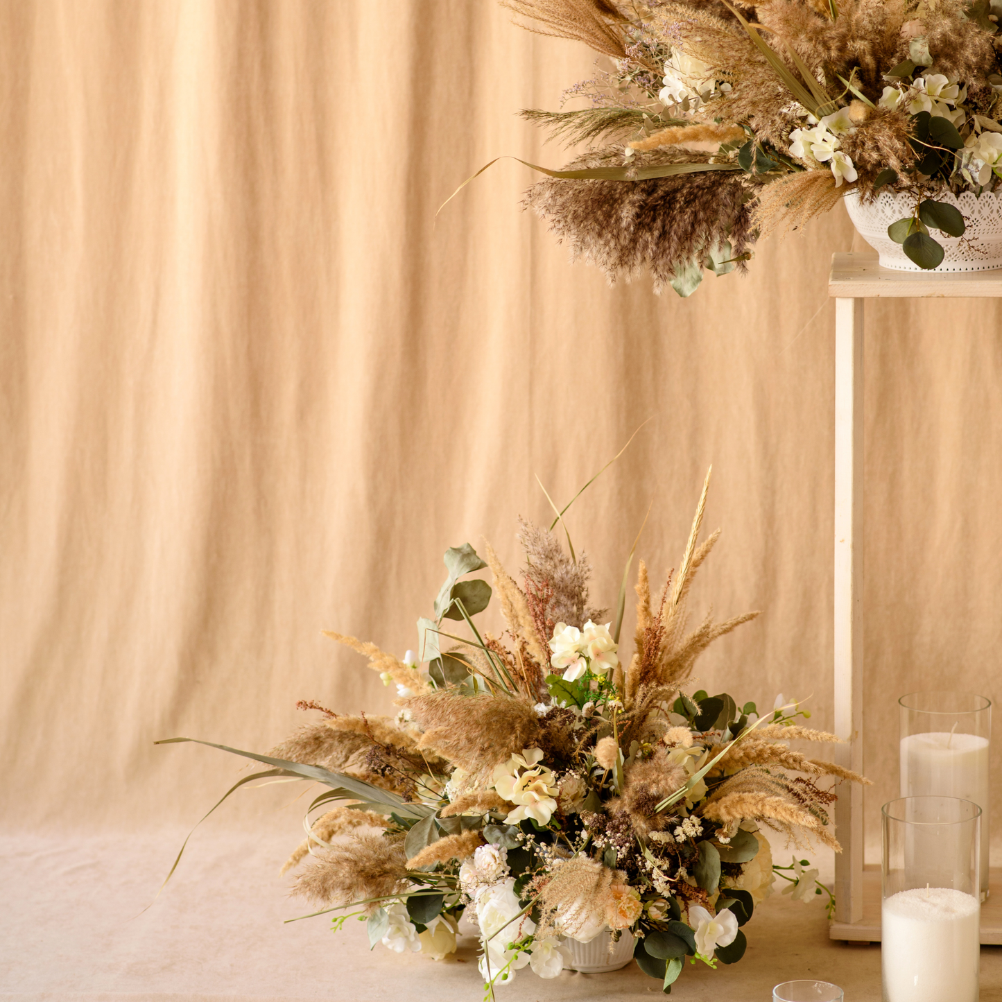 Floral arrangement with dried grasses, white flowers, and green foliage against a beige backdrop, with candles nearby. Prop hire, Prop hiring, Prop hire, event stylists Brisbane, brisbane event styling, brisbane event styling, event stylist Brisbane, Brisbane Wedding, Gold Coast Wedding, Floral Styling, Lush & Lavish Events