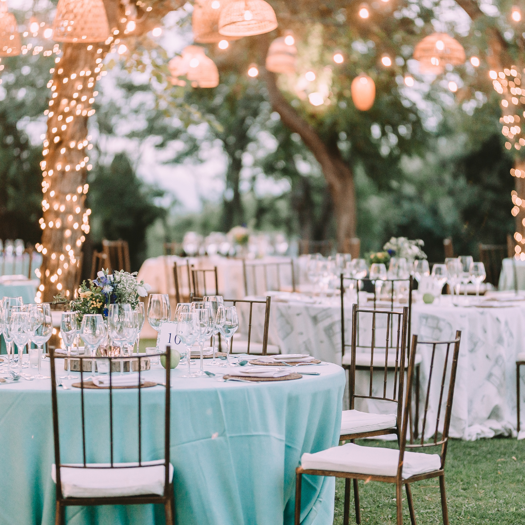 Outdoor event with round tables covered in light blue tablecloths, adorned with glassware, plates, and floral centerpieces. Metal chairs with white cushions surround the tables. String lights and lanterns hang from trees, creating a warm atmosphere. Prop hire, Prop hiring, Prop hire, event stylists Brisbane, brisbane event styling, brisbane event styling, event stylist Brisbane, Brisbane Wedding, Gold Coast Wedding, Floral Styling, Lush & Lavish Events