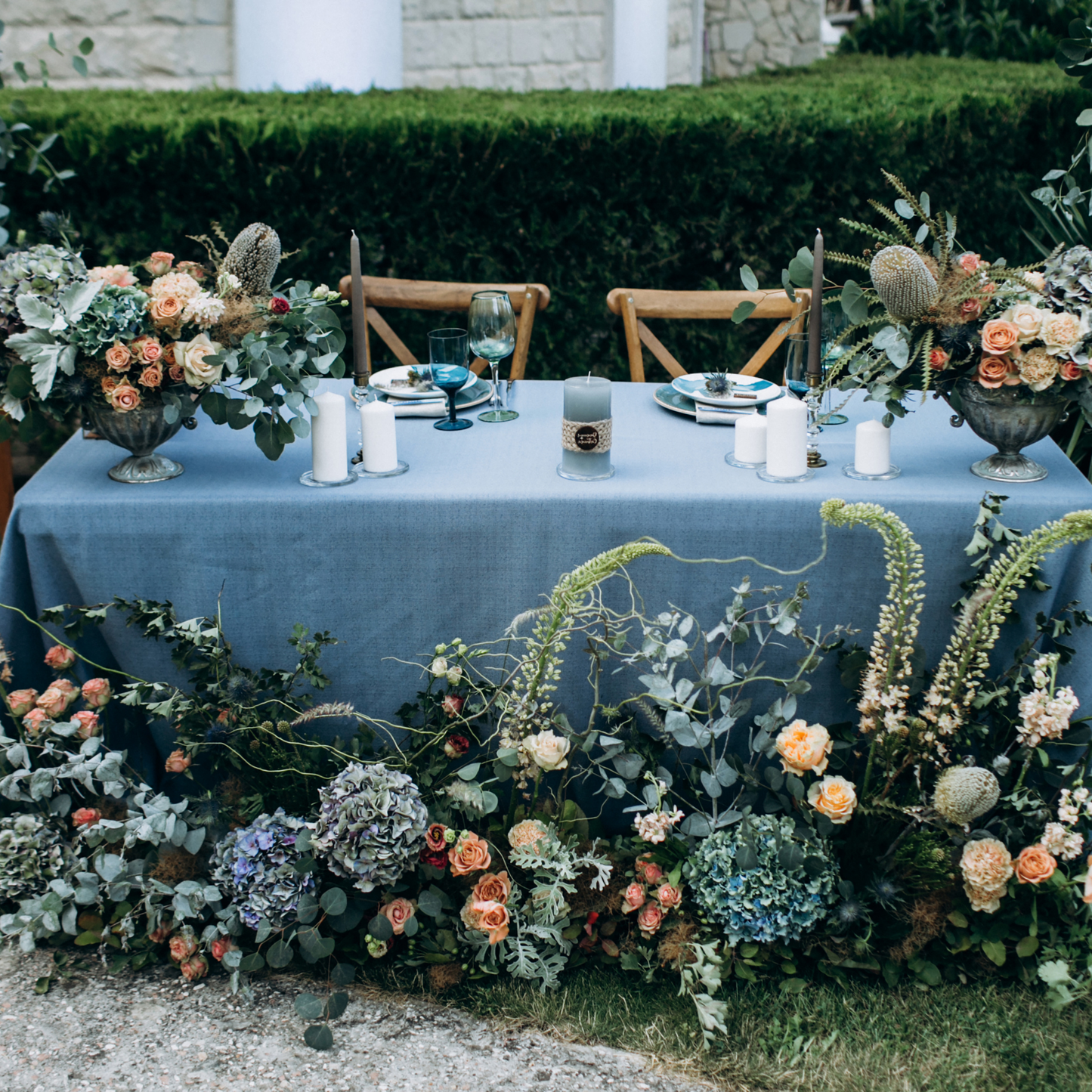 An outdoor table with a blue tablecloth, set with blue glasses, white plates, silver cutlery, white candles, and floral arrangements. The backdrop is a green hedge and a stone building with white columns. Prop hire, Prop hiring, Prop hire, event stylists Brisbane, brisbane event styling, brisbane event styling, event stylist Brisbane, Brisbane Wedding, Gold Coast Wedding, Floral Styling, Lush & Lavish Events