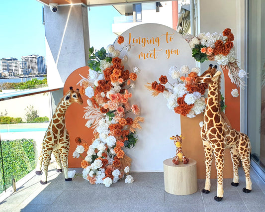 Two large giraffe statues and one small giraffe statue on a wooden stool. The backdrop includes a white and orange arch with orange and white flowers, with the text “longing to meet you.” Prop hire, Prop hiring, Prop hire, event stylists Brisbane, brisbane event styling, brisbane event styling, event stylist Brisbane, Brisbane Wedding, Gold Coast Wedding, Floral Styling, Lush & Lavish Events, Brisbane Tablescape, Gold Coast Tablescapes, Brisbane Candle Hire