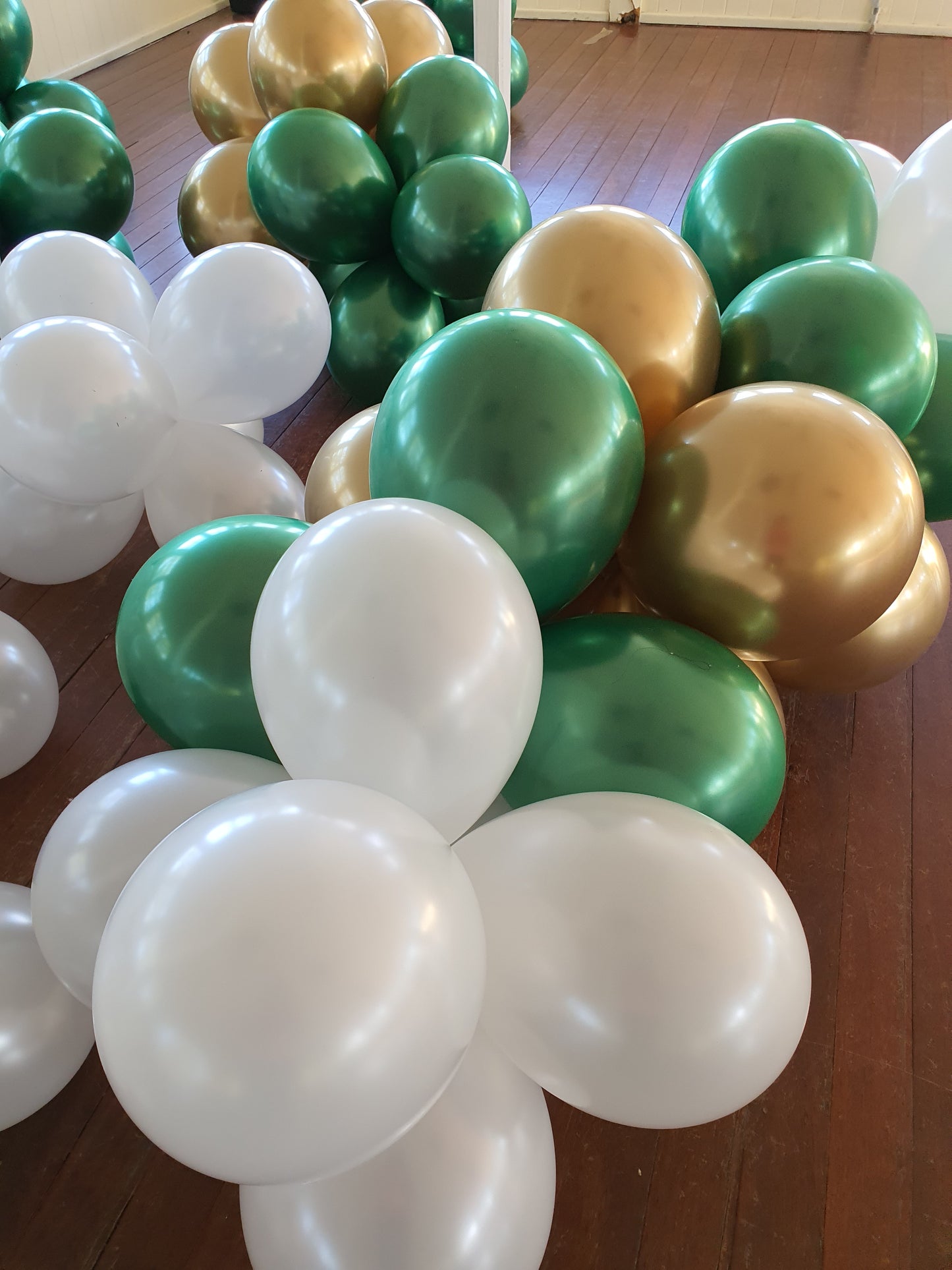 A cluster of white, green, and gold balloons arranged on a wooden floor, some forming flower-like shapes. Prop hire, Prop hiring, Prop hire, event stylists Brisbane, brisbane event styling, brisbane event styling, event stylist Brisbane, Brisbane Wedding, Gold Coast Wedding, Floral Styling, Lush & Lavish Events, Brisbane Corporate Styling, Gold Coast Corporate Styling, Brisbane Brand Activation
