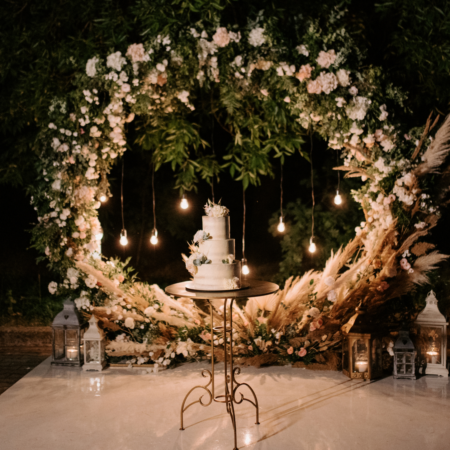 A round table with a cake in front of a circular floral arch adorned with white and pink flowers and greenery. Hanging lights illuminate the setup, with lanterns on the ground around the arch. Prop hire, Prop hiring, Prop hire, event stylists Brisbane, brisbane event styling, brisbane event styling, event stylist Brisbane, Brisbane Wedding, Gold Coast Wedding, Floral Styling, Lush & Lavish Events