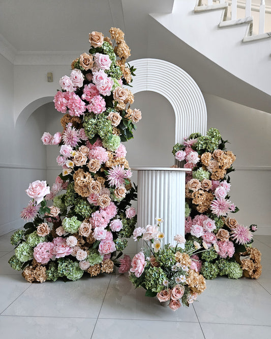 Pink, peach, and green floral arrangement around a white arch and pedestal, set against a white wall. Prop hire, Prop hiring, Prop hire, event stylists Brisbane, brisbane event styling, brisbane event styling, event stylist Brisbane, Brisbane Wedding, Gold Coast Wedding, Floral Styling, Lush & Lavish Events, Brisbane Tablescape, Gold Coast Tablescapes, Brisbane Candle Hire