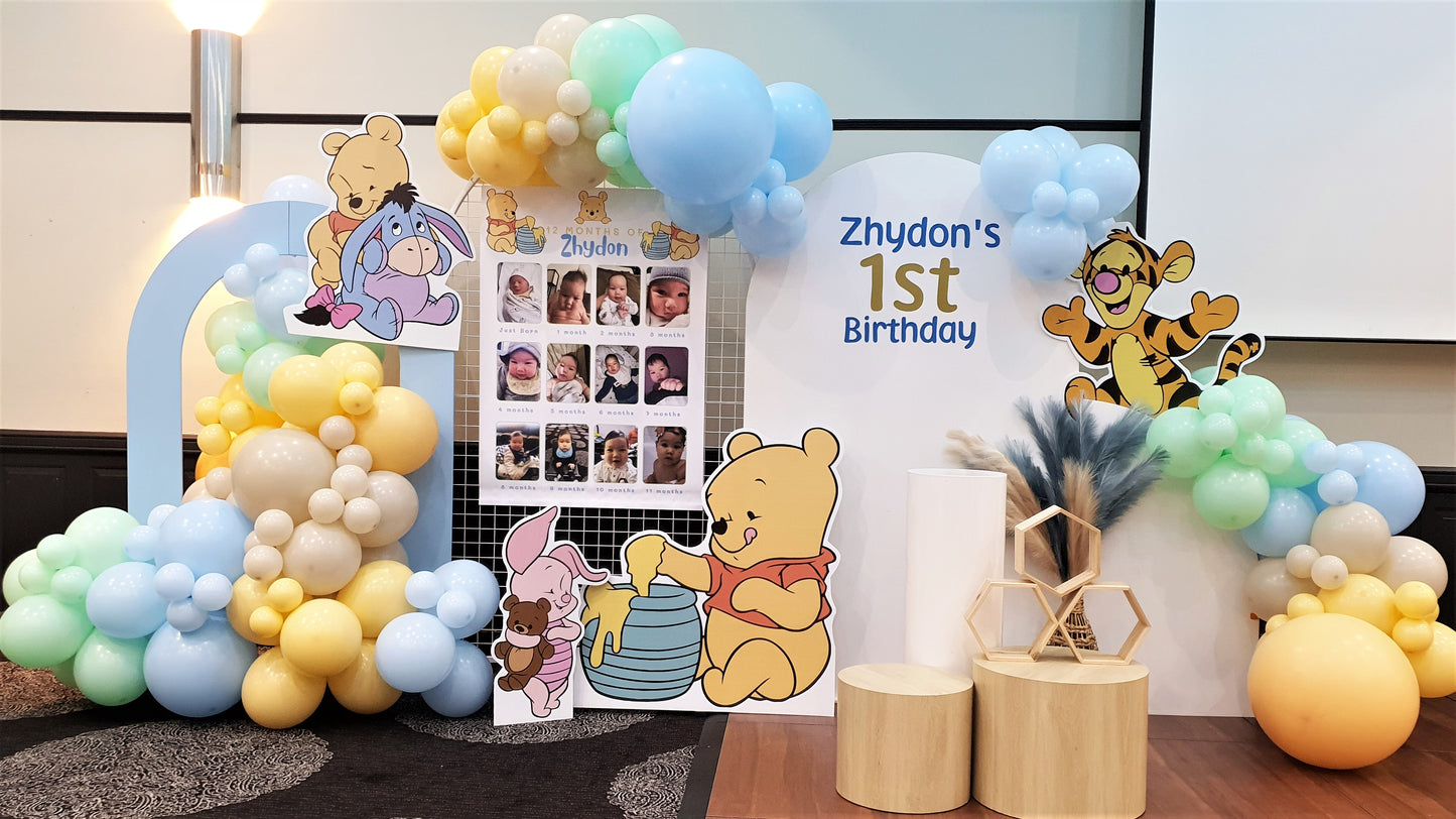 A Winnie the Pooh themed birthday party backdrop features a balloon arch in blue, yellow, and white. Cutouts of Winnie the Pooh characters, and a photo collage of the birthday child. Prop hire, Prop hiring, Prop hire, event stylists Brisbane, brisbane event styling, brisbane event styling, event stylist Brisbane, Brisbane Wedding, Gold Coast Wedding, Floral Styling, Lush & Lavish Events, Brisbane Tablescape, Gold Coast Tablescapes, Brisbane Candle Hire