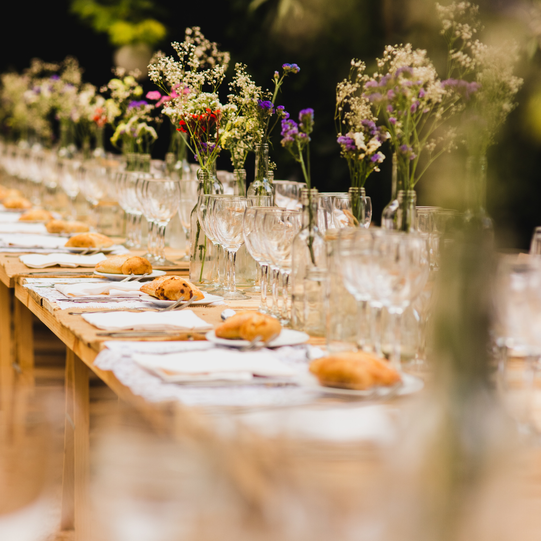 A long, elegantly set dining table adorned with white napkins, clear glassware, and small bread rolls on plates. Tall, slender vases with white baby’s breath and purple flowers decorate the table. The background is blurred greenery, suggesting an outdoor garden setting. Prop hire, Prop hiring, Prop hire, event stylists Brisbane, brisbane event styling, brisbane event styling, event stylist Brisbane, Brisbane Wedding, Gold Coast Wedding, Floral Styling, Lush & Lavish Events