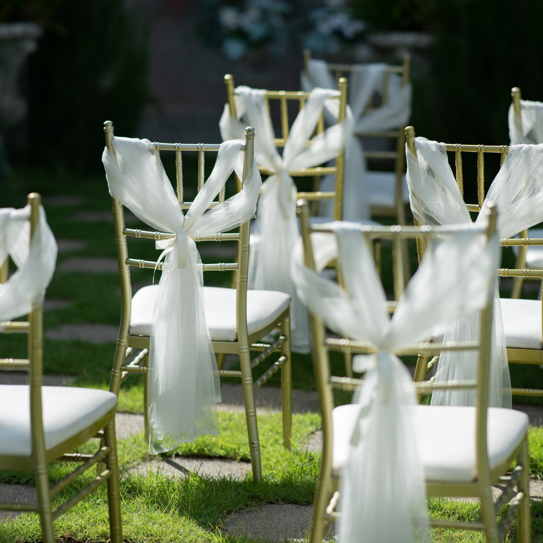Rows of gold-colored chairs with white cushions are arranged outdoors on a grassy area. Each chair is decorated with white, sheer fabric tied in bows on the backrests. Prop hire, Prop hiring, Prop hire, event stylists Brisbane, brisbane event styling, brisbane event styling, event stylist Brisbane, Brisbane Wedding, Gold Coast Wedding, Floral Styling, Lush & Lavish Events