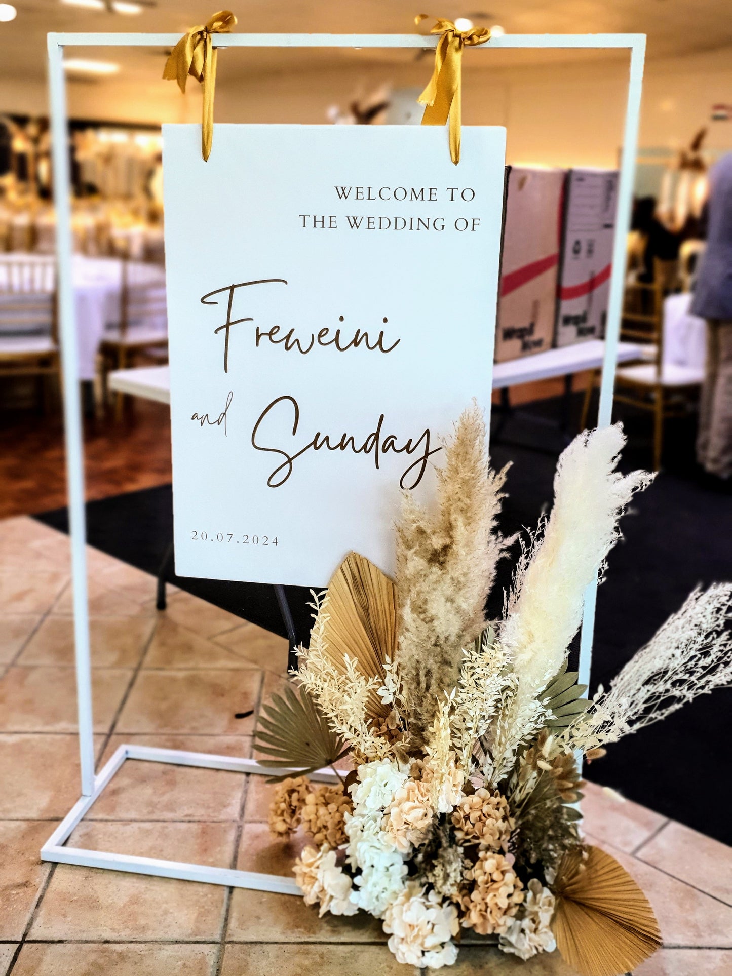 Wedding welcome sign with the text, "Welcome to the wedding of Freweini and Sunday, 24.07.2024," on a white stand with golden ribbons and dried flowers. Prop hire, Prop hiring, Prop hire, event stylists Brisbane, brisbane event styling, brisbane event styling, event stylist Brisbane, Brisbane Wedding, Gold Coast Wedding, Floral Styling, Lush & Lavish Events, Brisbane Tablescape, Gold Coast Tablescapes, Brisbane Candle Hire