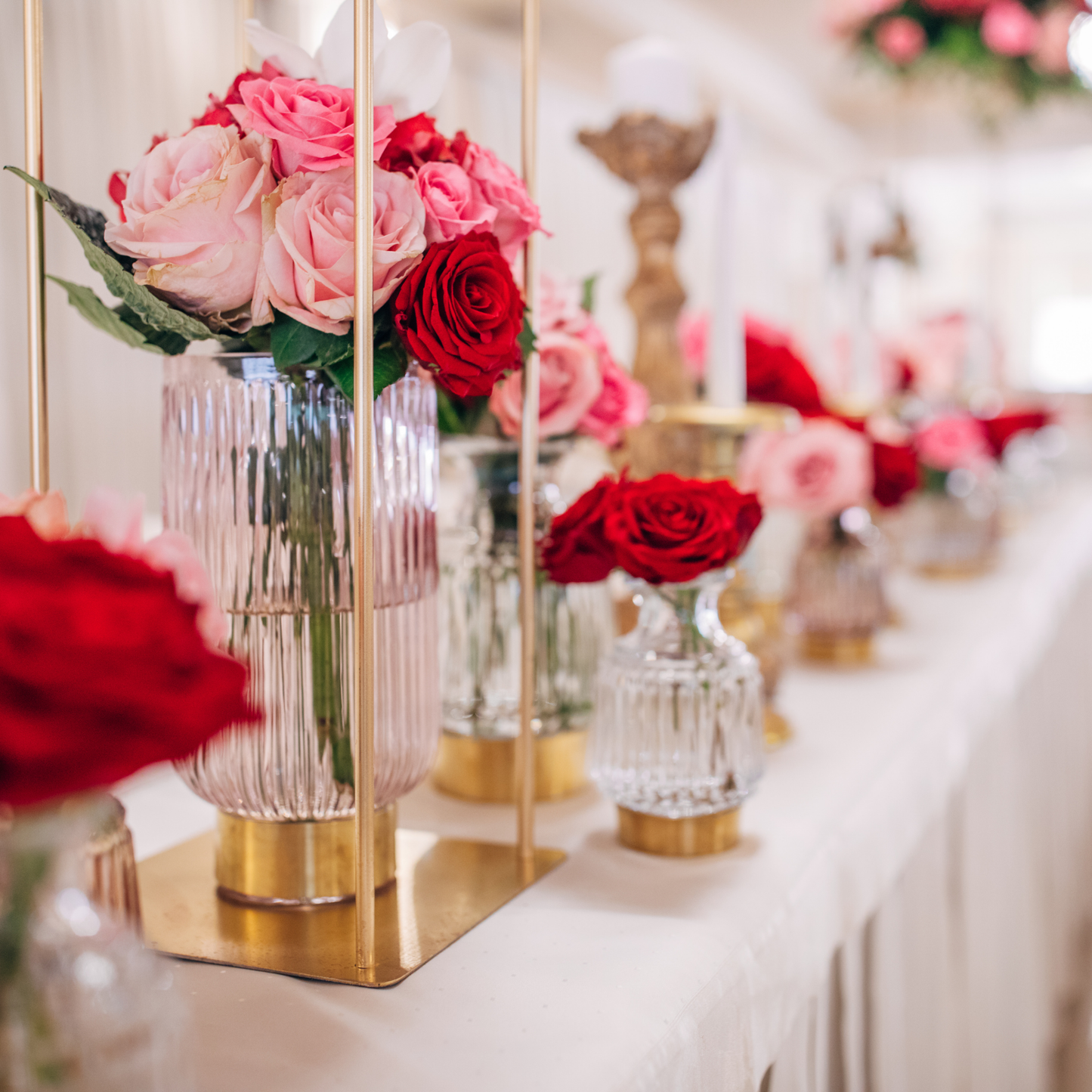 Decorative arrangement of vases filled with red and pink roses on a white tablecloth with gold stands, set against a softly blurred background. Prop hire, Prop hiring, Prop hire, event stylists Brisbane, brisbane event styling, brisbane event styling, event stylist Brisbane, Brisbane Wedding, Gold Coast Wedding, Floral Styling, Lush & Lavish Events, Brisbane Tablescape, Gold Coast Tablescapes, Brisbane Candle Hire
