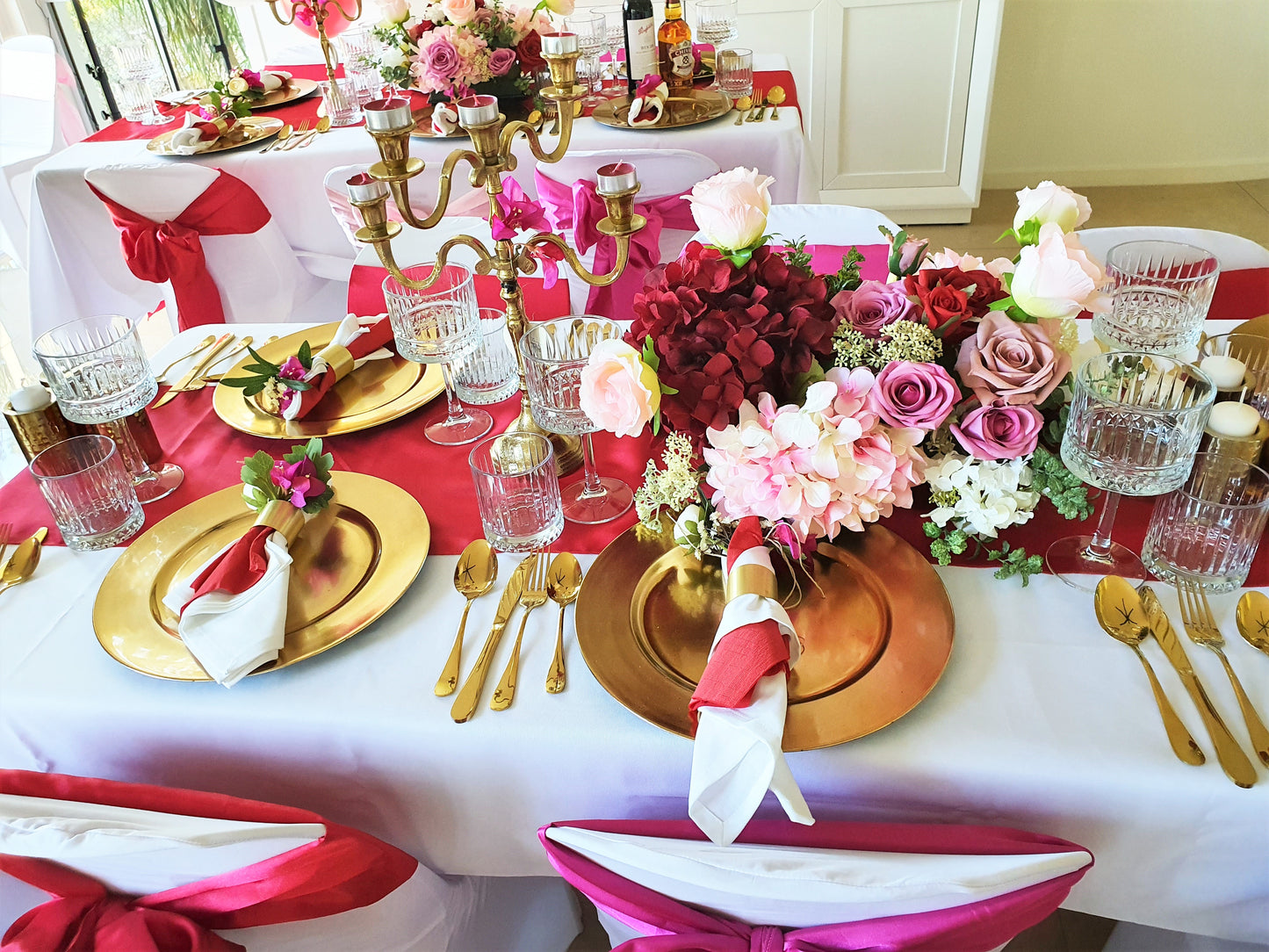 A long rectangular table with a white tablecloth set for a special occasion, decorated with gold chargers, red napkins, and a large centerpiece of pink and red flowers. Prop hire, Prop hiring, Prop hire, event stylists Brisbane, brisbane event styling, brisbane event styling, event stylist Brisbane, Brisbane Wedding, Gold Coast Wedding, Floral Styling, Lush & Lavish Events, Brisbane Tablescape, Gold Coast Tablescapes, Brisbane Candle Hire