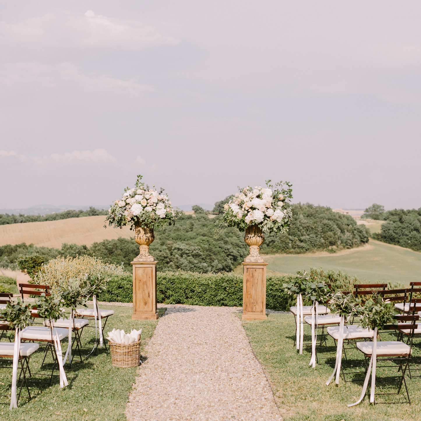 Outdoor wedding with a gravel aisle, white chairs with greenery, and floral arrangements on pedestals. Rolling hills and trees in the background. Prop hire, Prop hiring, Prop hire, event stylists Brisbane, brisbane event styling, brisbane event styling, event stylist Brisbane, Brisbane Wedding, Gold Coast Wedding, Floral Styling, Lush & Lavish Events