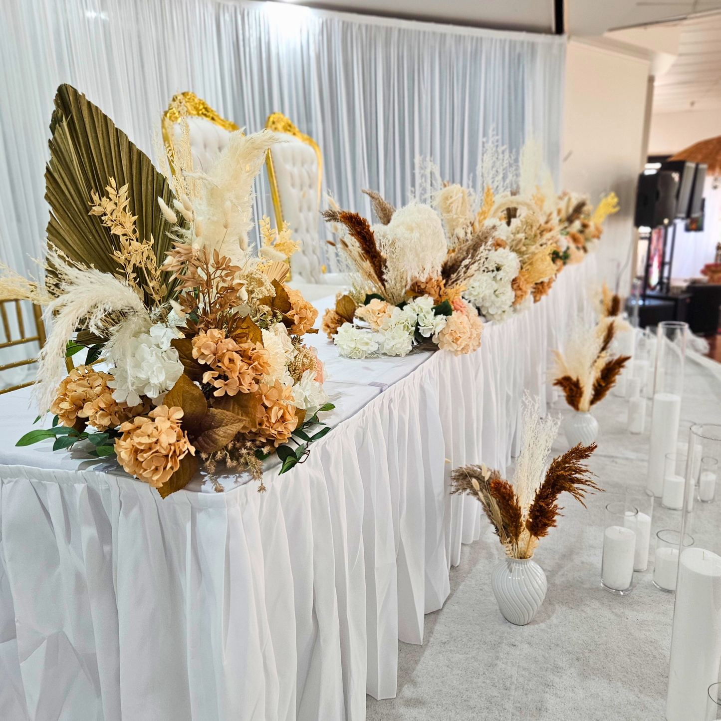 A wedding reception bridal table features a stunning floral centerpiece in white, cream, and peach tones with gold and green foliage. The table is set against a sheer white curtain backdrop, with white vases and cylindrical candles adding to the elegant atmosphere. Prop hire, Prop hiring, Prop hire, event stylists Brisbane, brisbane event styling, brisbane event styling, event stylist Brisbane, Brisbane Wedding, Gold Coast Wedding, Floral Styling, Lush & Lavish Events, Brisbane Tablescape, Gold Coast Tables