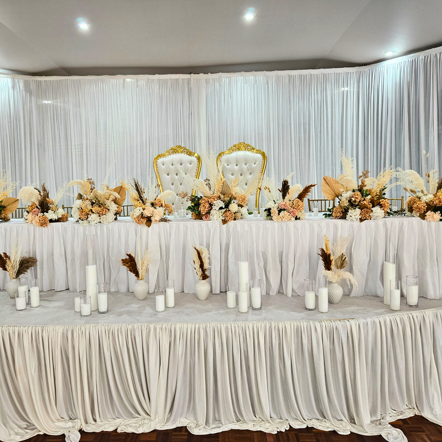 Wedding reception head table with white draped tablecloth, ornate white chairs, peach and white flowers, green foliage, tall white candles, and a sheer white curtain backdrop. Prop hire, Prop hiring, Prop hire, event stylists Brisbane, brisbane event styling, brisbane event styling, event stylist Brisbane, Brisbane Wedding, Gold Coast Wedding, Floral Styling, Lush & Lavish Events, Brisbane Tablescape, Gold Coast Tablescapes, Brisbane Candle Hire