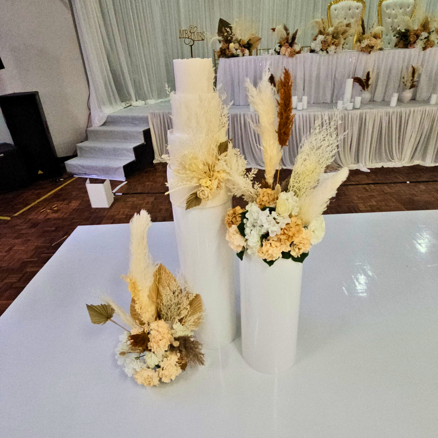 Cake stand with two white pillars of different heights adorned with dried flowers in white, cream, and beige. Background shows decorated chairs and a stage, indicating a celebration. Prop hire, Prop hiring, Prop hire, event stylists Brisbane, brisbane event styling, brisbane event styling, event stylist Brisbane, Brisbane Wedding, Gold Coast Wedding, Floral Styling, Lush & Lavish Events, Brisbane Tablescape, Gold Coast Tablescapes, Brisbane Candle Hire