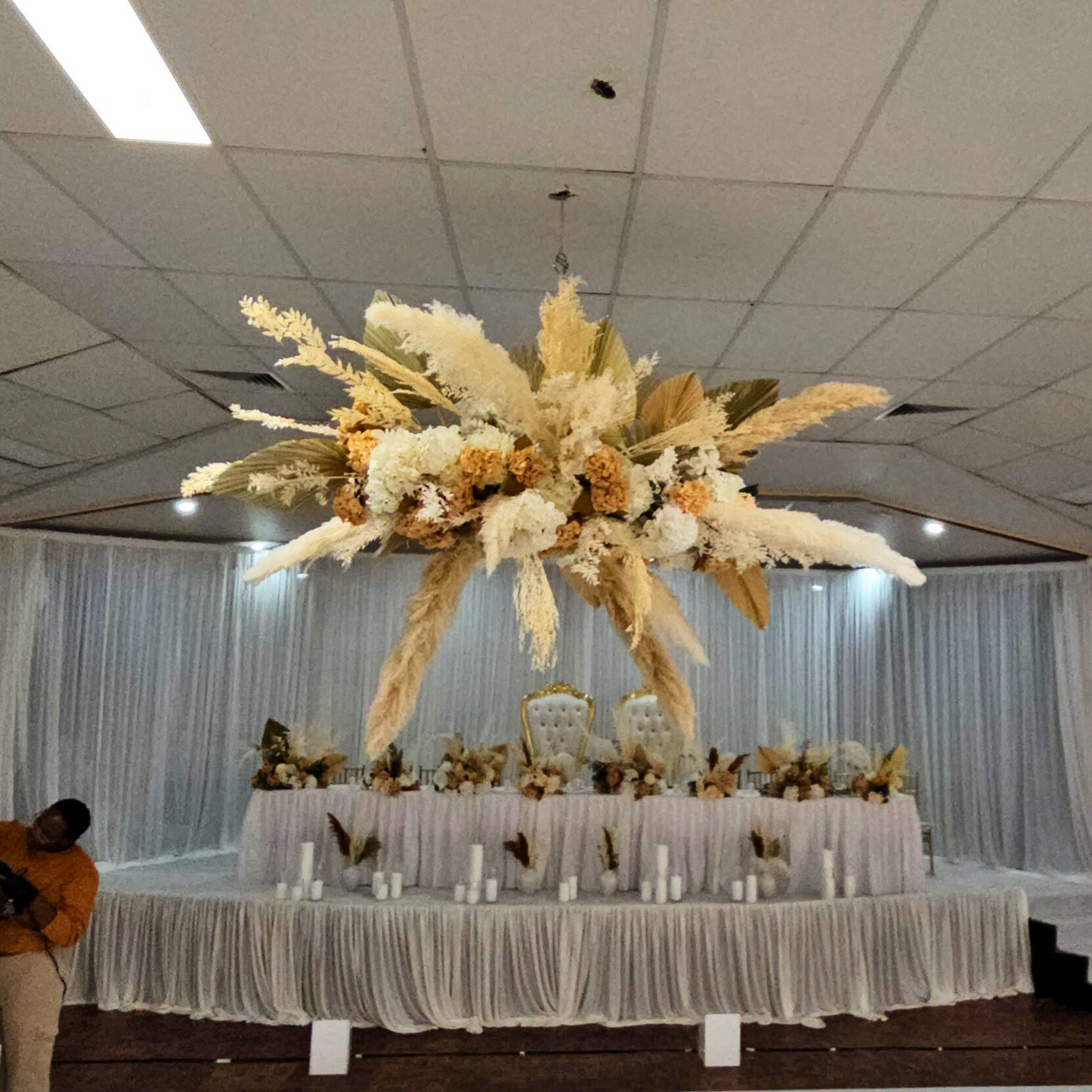 Ornate dried floral arrangement with white and pale yellow flowers, pampas grass, and foliage, suspended above a decorated table against a white backdrop in an event hall. Prop hire, Prop hiring, Prop hire, event stylists Brisbane, brisbane event styling, brisbane event styling, event stylist Brisbane, Brisbane Wedding, Gold Coast Wedding, Floral Styling, Lush & Lavish Events, Brisbane Tablescape, Gold Coast Tablescapes, Brisbane Candle Hire