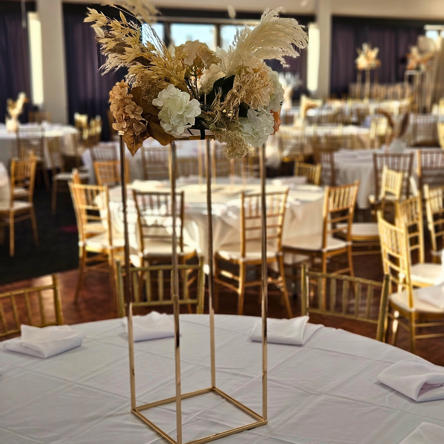 Wedding reception venue with round tables covered in white tablecloths, gold Chiavari chairs, and tall centerpieces with white and beige flowers on geometric gold stands. Prop hire, Prop hiring, Prop hire, event stylists Brisbane, brisbane event styling, brisbane event styling, event stylist Brisbane, Brisbane Wedding, Gold Coast Wedding, Floral Styling, Lush & Lavish Events, Brisbane Tablescape, Gold Coast Tablescapes, Brisbane Candle Hire