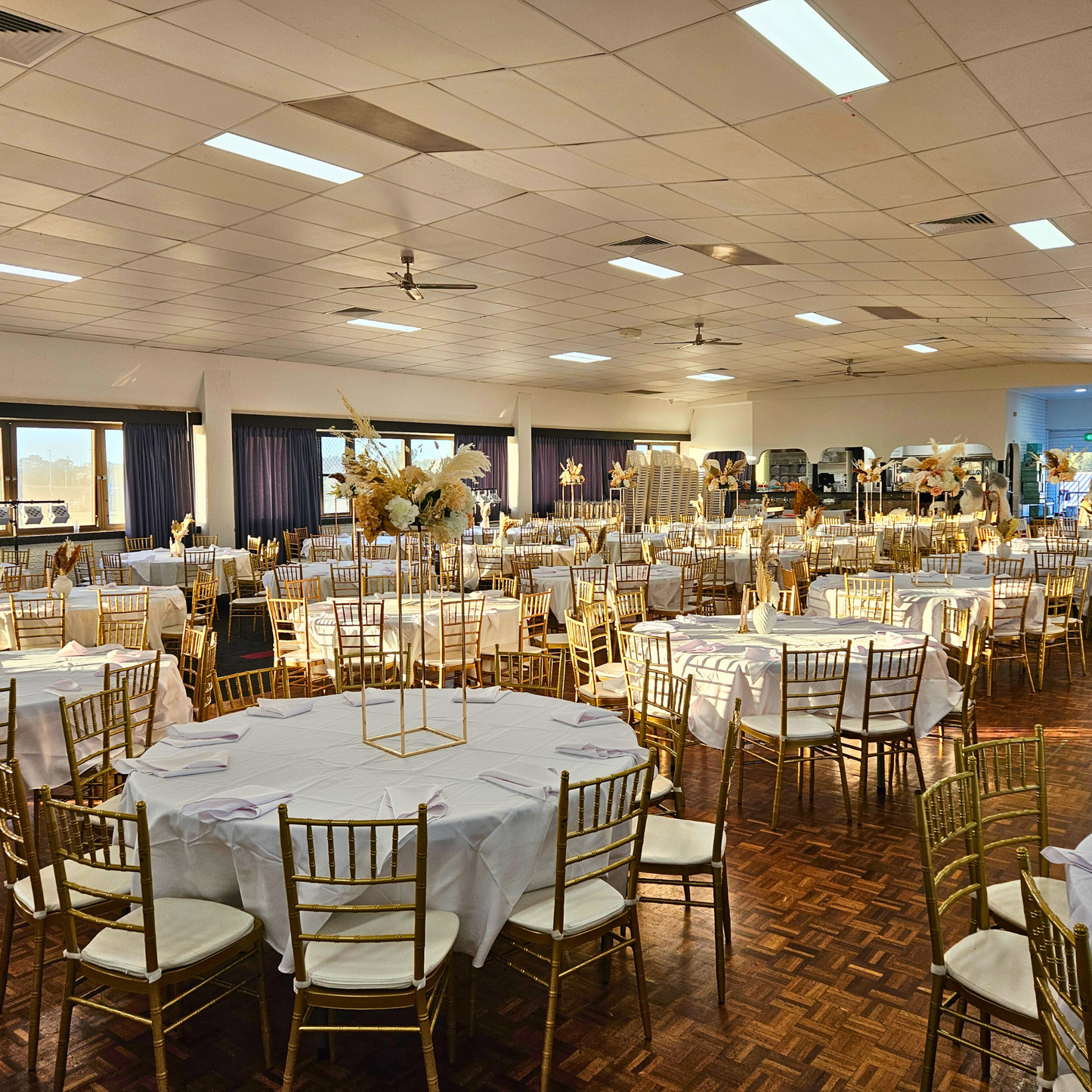 Wedding reception hall with round tables, white linens, golden chairs, parquet floor, recessed lighting, large windows with dark curtains, and a floral arrangement, creating a formal and festive ambiance. Prop hire, Prop hiring, Prop hire, event stylists Brisbane, brisbane event styling, brisbane event styling, event stylist Brisbane, Brisbane Wedding, Gold Coast Wedding, Floral Styling, Lush & Lavish Events, Brisbane Tablescape, Gold Coast Tablescapes, Brisbane Candle Hire