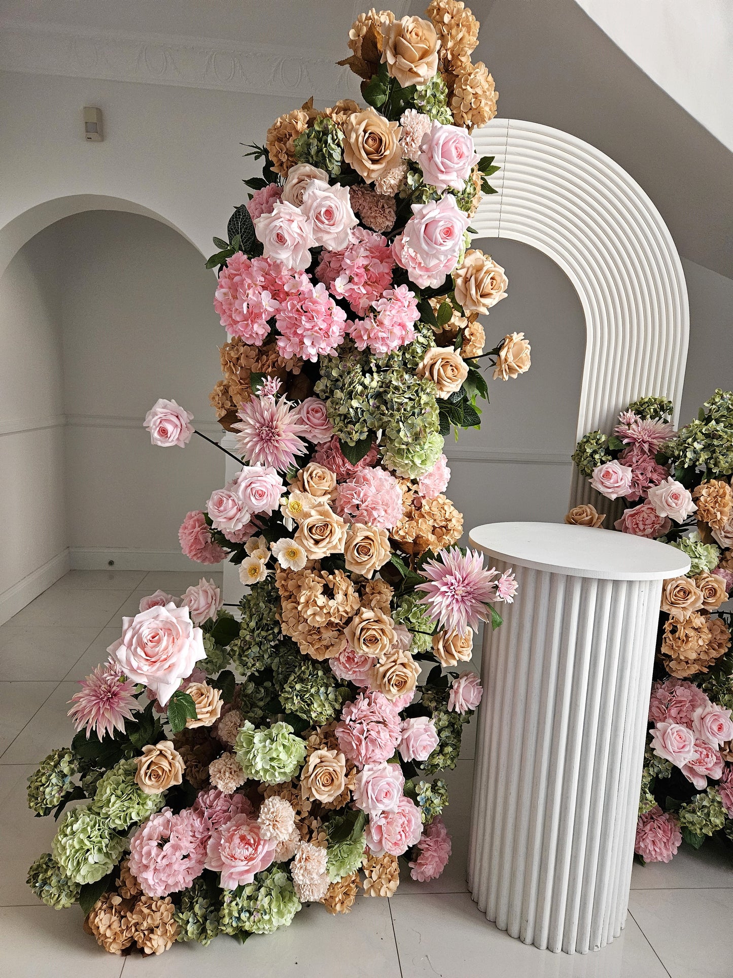 Floral arrangement with pink roses, beige roses, green hydrangeas, and pink chrysanthemums beside a white pedestal and arch, against a white wall. Prop hire, Prop hiring, Prop hire, event stylists Brisbane, brisbane event styling, brisbane event styling, event stylist Brisbane, Brisbane Wedding, Gold Coast Wedding, Floral Styling, Lush & Lavish Events, Brisbane Tablescape, Gold Coast Tablescapes, Brisbane Candle Hire
