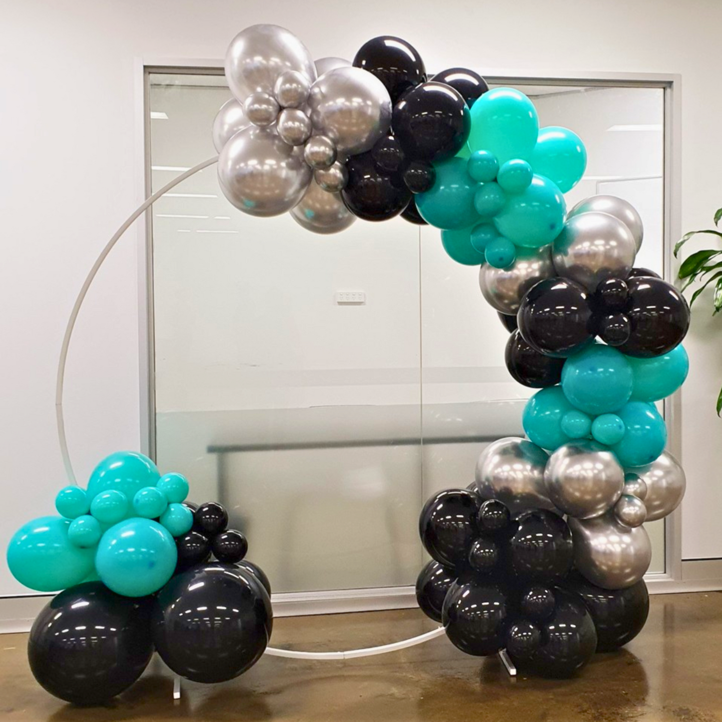 A decorative balloon arch in black, silver, and teal near a glass door in a room with white walls and a light floor. Larger balloons are at the base, transitioning to smaller ones at the top. Prop hire, Prop hiring, Prop hire, event stylists Brisbane, brisbane event styling, brisbane event styling, event stylist Brisbane, Brisbane Wedding, Gold Coast Wedding, Floral Styling, Lush & Lavish Events, Brisbane Corporate Styling, Gold Coast Corporate Styling, Brisbane Brand Activation