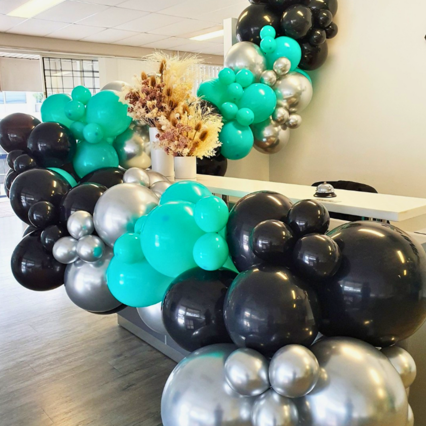 A festive balloon arch in shades of green, black, and silver decorates an indoor space. Brisbane corporate balloon garland, Gold Coast corporate balloon garland, Brisbane brand activation balloons, Gold Coast brand activation balloons, Brisbane corporate event balloons, Gold Coast corporate event balloons, Brisbane product launch balloons, Gold Coast product launch balloons, Brisbane grand opening balloons, Gold Coast grand opening balloons, Brisbane corporate event styling, Gold Coast corporate event styli