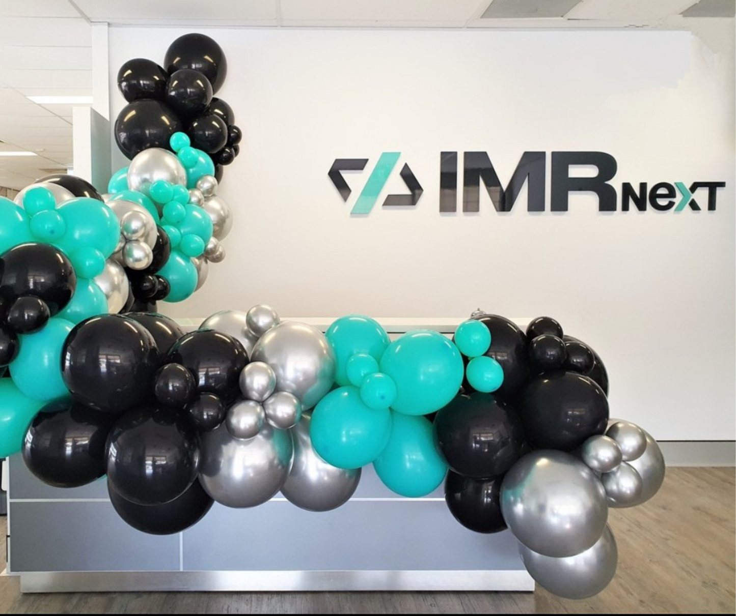 A reception area features a desk with the IMRneXt logo and a wall decorated with the same logo and clusters of black, dark blue, light blue, and silver balloons. Prop hire, Prop hiring, Prop hire, event stylists Brisbane, brisbane event styling, brisbane event styling, event stylist Brisbane, Brisbane Wedding, Gold Coast Wedding, Floral Styling, Lush & Lavish Events, Brisbane Corporate Styling, Gold Coast Corporate Styling, Brisbane Brand Activation