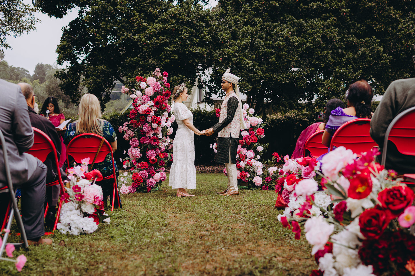 Outdoor wedding ceremony with a couple standing in front of a floral arch decorated with pink and red flowers. Guests are seated on red chairs in a garden setting. Prop hire, Prop hiring, Prop hire, event stylists Brisbane, brisbane event styling, brisbane event styling, event stylist Brisbane, Brisbane Wedding, Gold Coast Wedding, Floral Styling, Lush & Lavish Events, Brisbane Corporate Styling, Gold Coast Corporate Styling, Brisbane Brand Activation