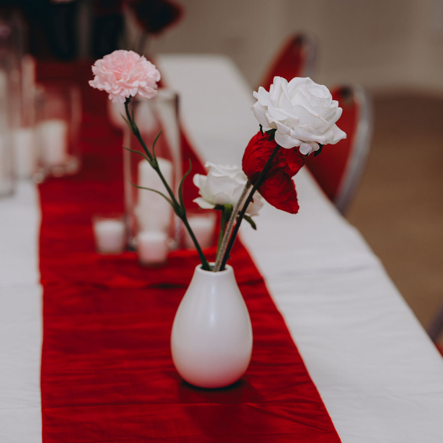 White vase with pink and white flowers on a table with a red runner, blurred candles, and chairs in the background. Prop hire, Prop hiring, Prop hire, event stylists Brisbane, brisbane event styling, brisbane event styling, event stylist Brisbane, Brisbane Wedding, Gold Coast Wedding, Floral Styling, Lush & Lavish Events, Brisbane Corporate Styling, Gold Coast Corporate Styling, Brisbane Brand Activation