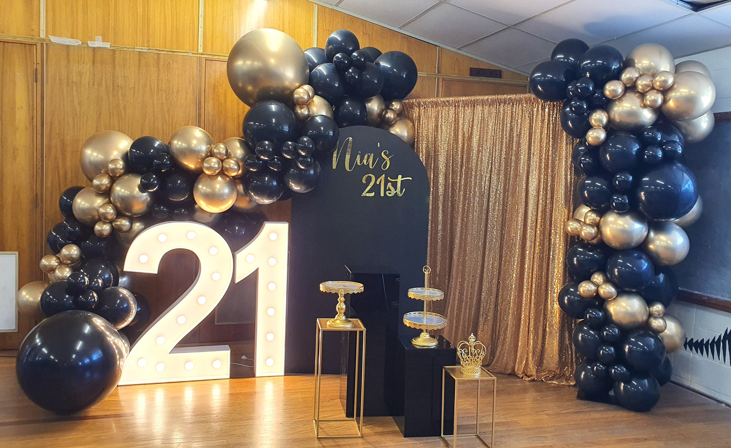 A decorated event space for a 21st birthday celebration. The setup includes a large illuminated number "21" on the left, surrounded by black and gold balloons. In the center, there is a black stand with the text "Elias's 21st" written in gold. To the right, there is a gold sequin curtain backdrop with more black and gold balloons. The floor has a wooden finish, and there are two small tables with gold decorations in front of the stand. Prop hire, Prop hiring, Prop hire, event stylists Brisbane, brisbane eve
