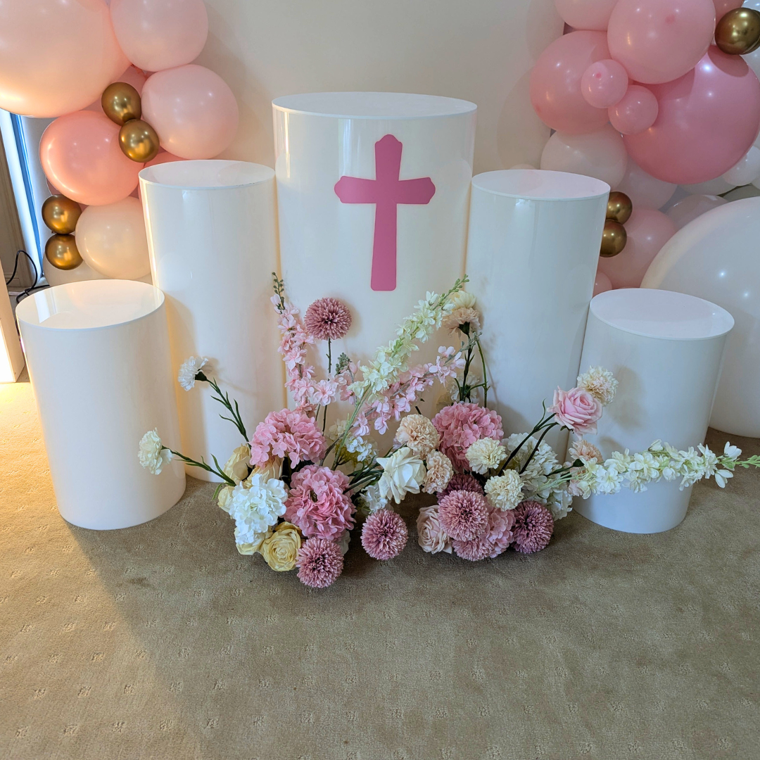 Decorative setup with white plinth, pink flowers, and a candle with a cross symbol against a balloon arch. Prop hire, Prop hiring, Prop hire, event stylists Brisbane, brisbane event styling, brisbane event styling, event stylist Brisbane, Brisbane Wedding, Gold Coast Wedding, Floral Styling, Lush & Lavish Events, Brisbane Corporate Styling, Gold Coast Corporate Styling, Brisbane Brand Activation, Gold Coast Tablescapes, Brisbane Candle Hire