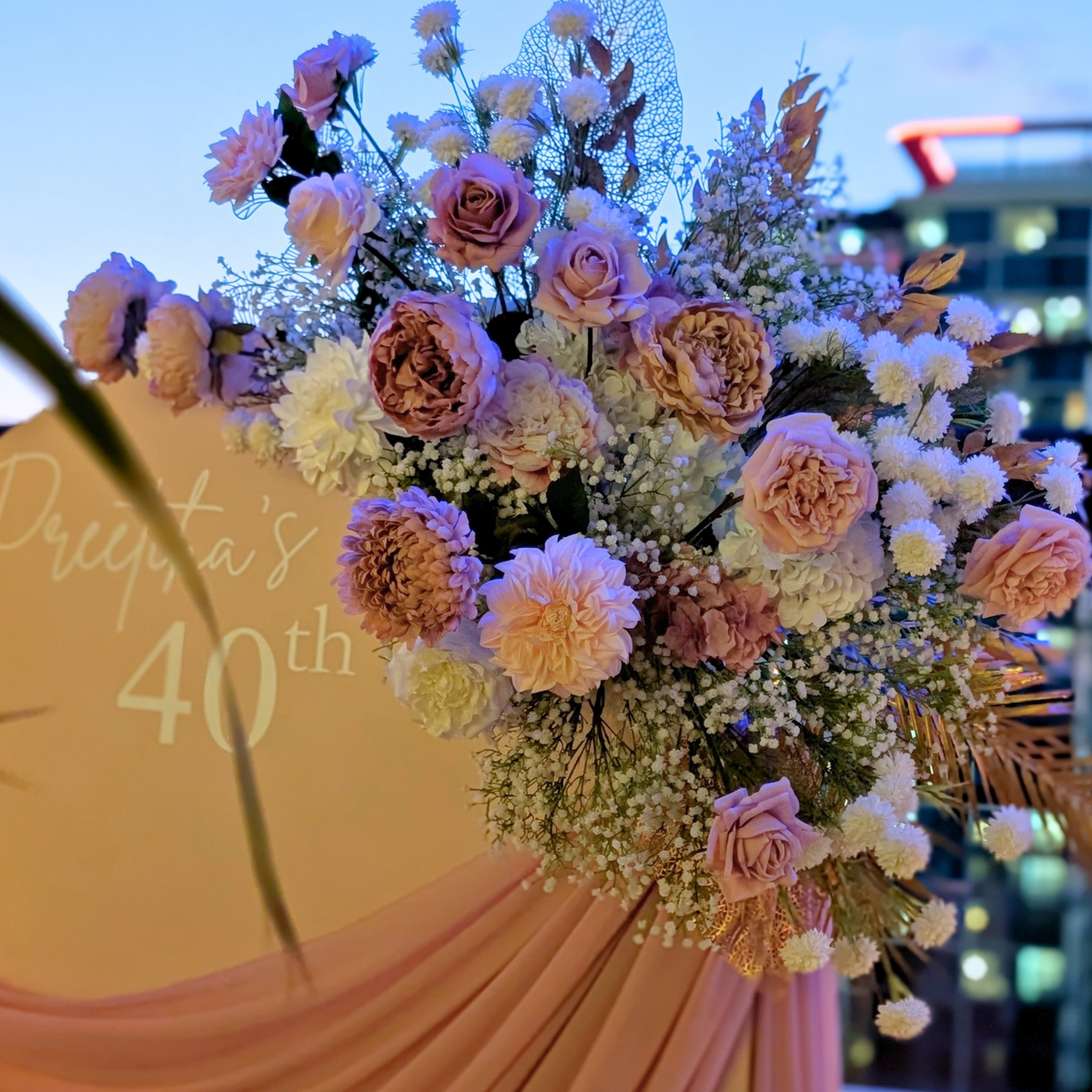 Outdoor wedding-style event backdrop with a cream arched board reading 'Preetika's 40th,' draped blush pink fabric, large floral arrangements in pinks and white, and a city skyline at dusk in the background. Styled at Maya Rooftop bar Brisbane by Lush and Lavish Event an event styling, event planning and on-the-day coordination business.
