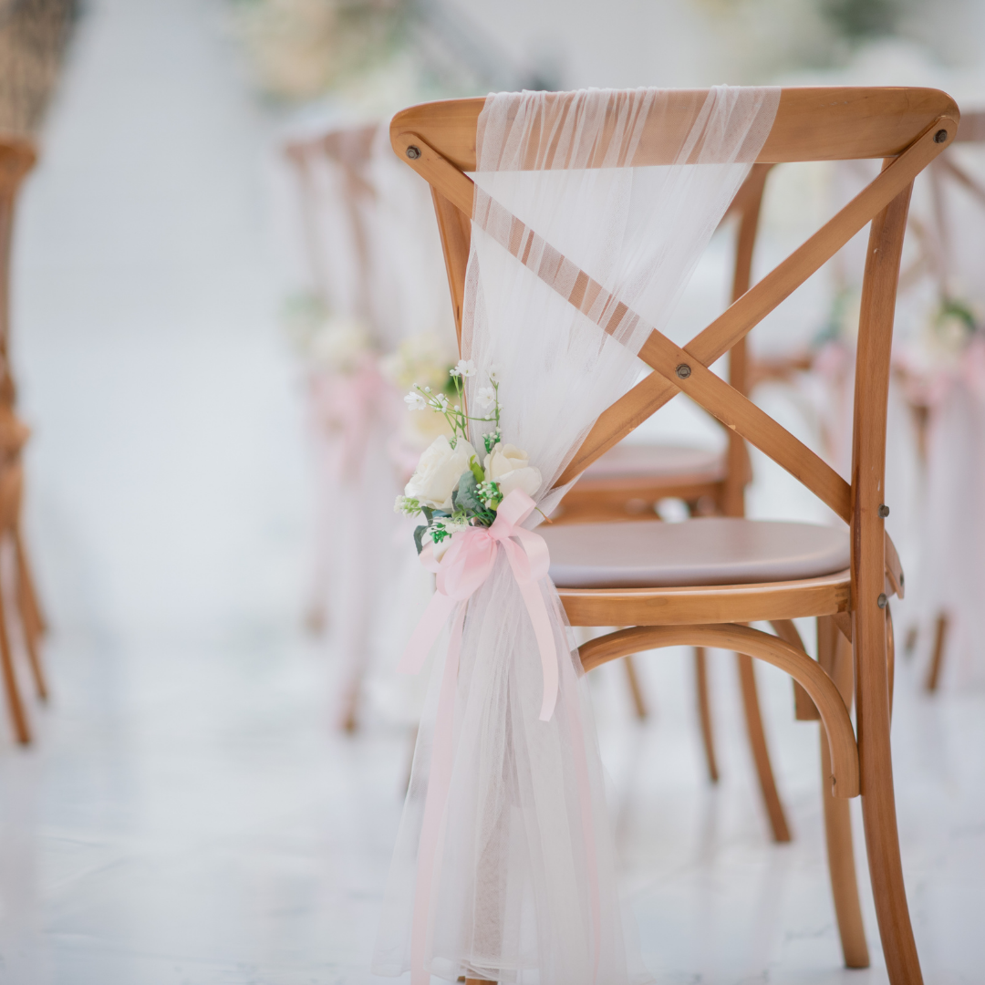 A wooden chair with a cross-back design is decorated with white tulle fabric draped over the backrest. The tulle is gathered and tied with a pink ribbon, which also holds a small bouquet of white and pink flowers. Prop hire, Prop hiring, Prop hire, event stylists Brisbane, brisbane event styling, brisbane event styling, event stylist Brisbane, Brisbane Wedding, Gold Coast Wedding, Floral Styling, Lush & Lavish Events