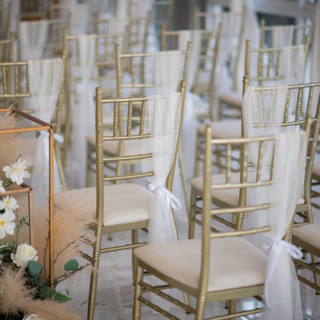 Rows of gold-colored chairs with white cushions are arranged in a venue. Each chair is adorned with a white sheer fabric tied in a bow at the back. There are floral decorations with white flowers and greenery visible in the foreground. Prop hire, Prop hiring, Prop hire, event stylists Brisbane, brisbane event styling, brisbane event styling, event stylist Brisbane, Brisbane Wedding, Gold Coast Wedding, Floral Styling, Lush & Lavish Events