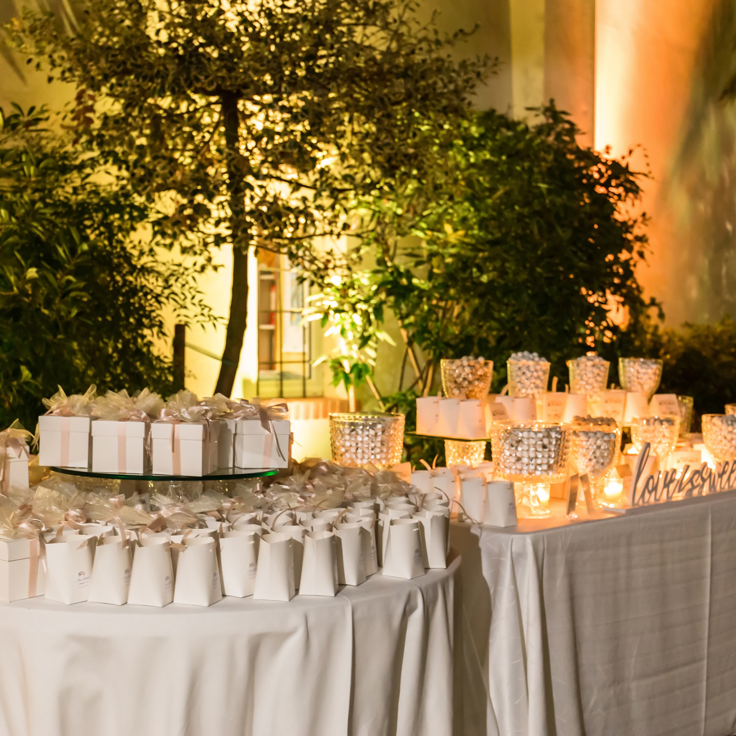 Elegantly decorated table with white gift boxes and glass containers filled with white candies, set against a lush greenery backdrop and warm lighting. Prop hire, Prop hiring, Prop hire, event stylists Brisbane, brisbane event styling, brisbane event styling, event stylist Brisbane, Brisbane Wedding, Gold Coast Wedding, Floral Styling, Lush & Lavish Events, Brisbane Tablescape, Gold Coast Tablescapes, Brisbane Candle Hire