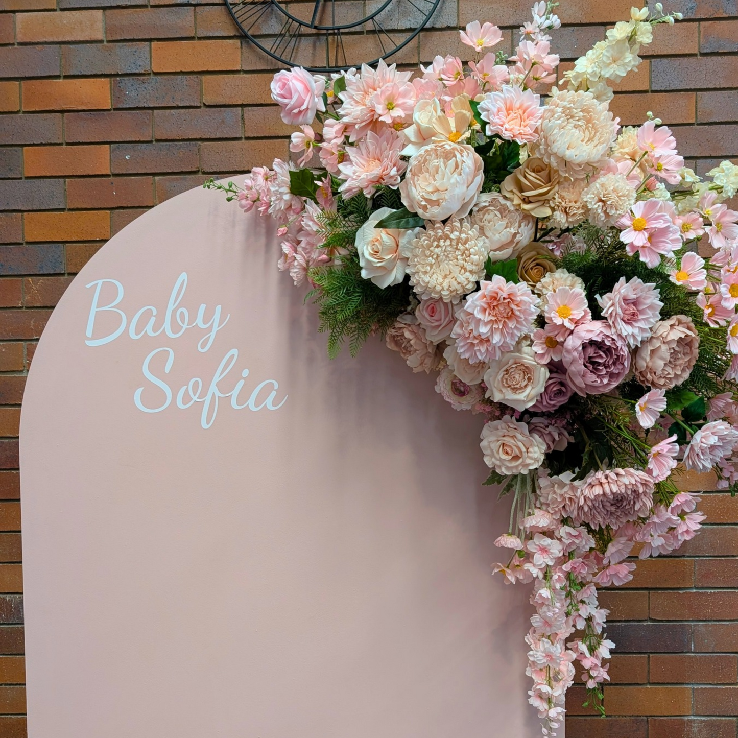 A baby shower backdrop setup featuring a tall, light pink arch with the text 'Baby Sofia' in white script, decorated with a cascading floral arrangement of pink and cream roses, set against a rustic brick wall with a white acrylic plinth. Styled at Future Brewing Woolloongabba by Lush and Lavish Events a Brisbane and Gold coast based Event styling, event planning, on-the-day coordination and Event MC services business.