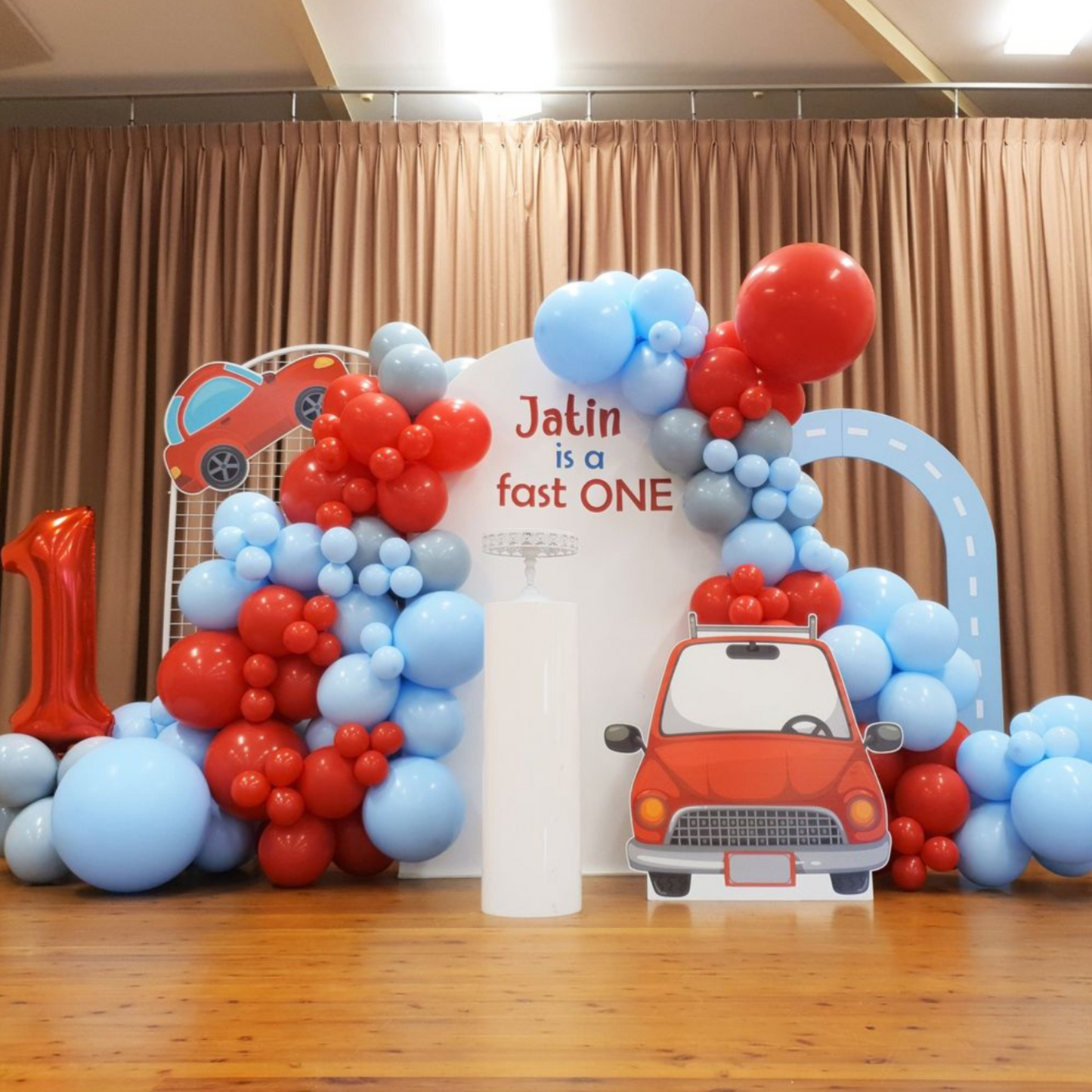Vibrant first birthday party setup with a large number one-shaped balloon arrangement in blue and red, featuring a cars and racing theme. Backdrop displays ‘Jatin is a fast ONE’ with playful font, accompanied by balloon clusters resembling traffic lights and car shapes, set in an indoor venue with curtains. Lush and Lavish Events