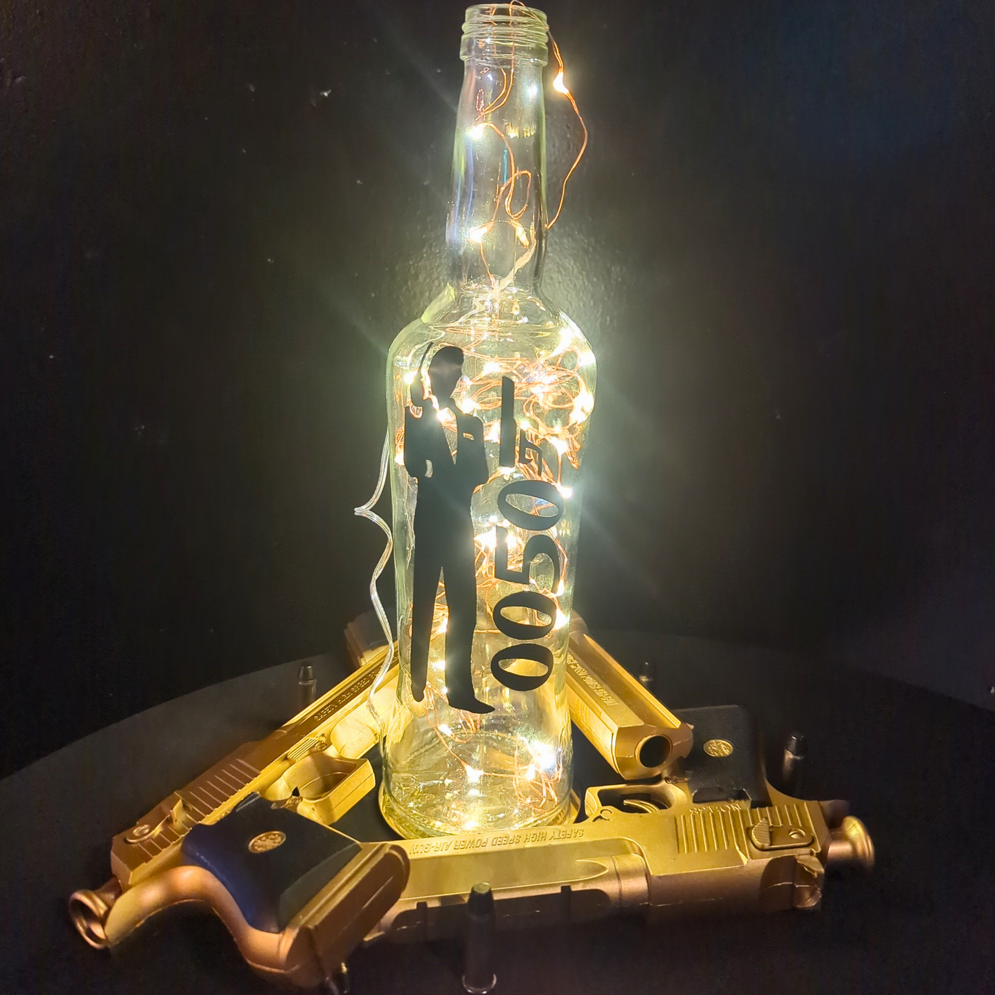 A glass bottle with illuminated light bulbs inside sits on a dark surface. Two golden toy guns form an X beneath it. Inside the bottle, a silhouette of James Bond stands next to floating numbers '0050'. The black background enhances the glow and contrast. Prop hire, Prop hiring, Prop hire, event stylists Brisbane, brisbane event styling, brisbane event styling, event stylist Brisbane, Brisbane Wedding, Gold Coast Wedding, Floral Styling, Lush & Lavish Events