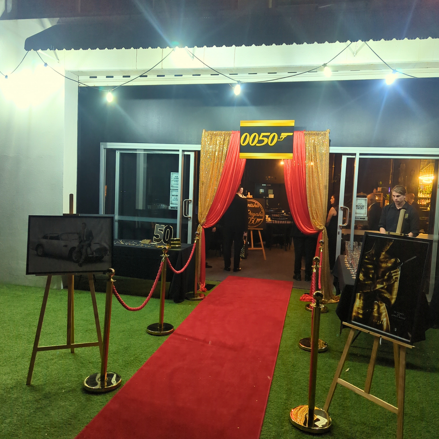 An entrance adorned with a red carpet, gold curtains, and stanchions leads to a doorway marked "0050." Overhead string lights illuminate the scene. Prop hire, Prop hiring, Prop hire, event stylists Brisbane, brisbane event styling, brisbane event styling, event stylist Brisbane, Brisbane Wedding, Gold Coast Wedding, Floral Styling, Lush & Lavish Events, Brisbane Tablescape, Gold Coast Tablescapes, Brisbane Candle Hire