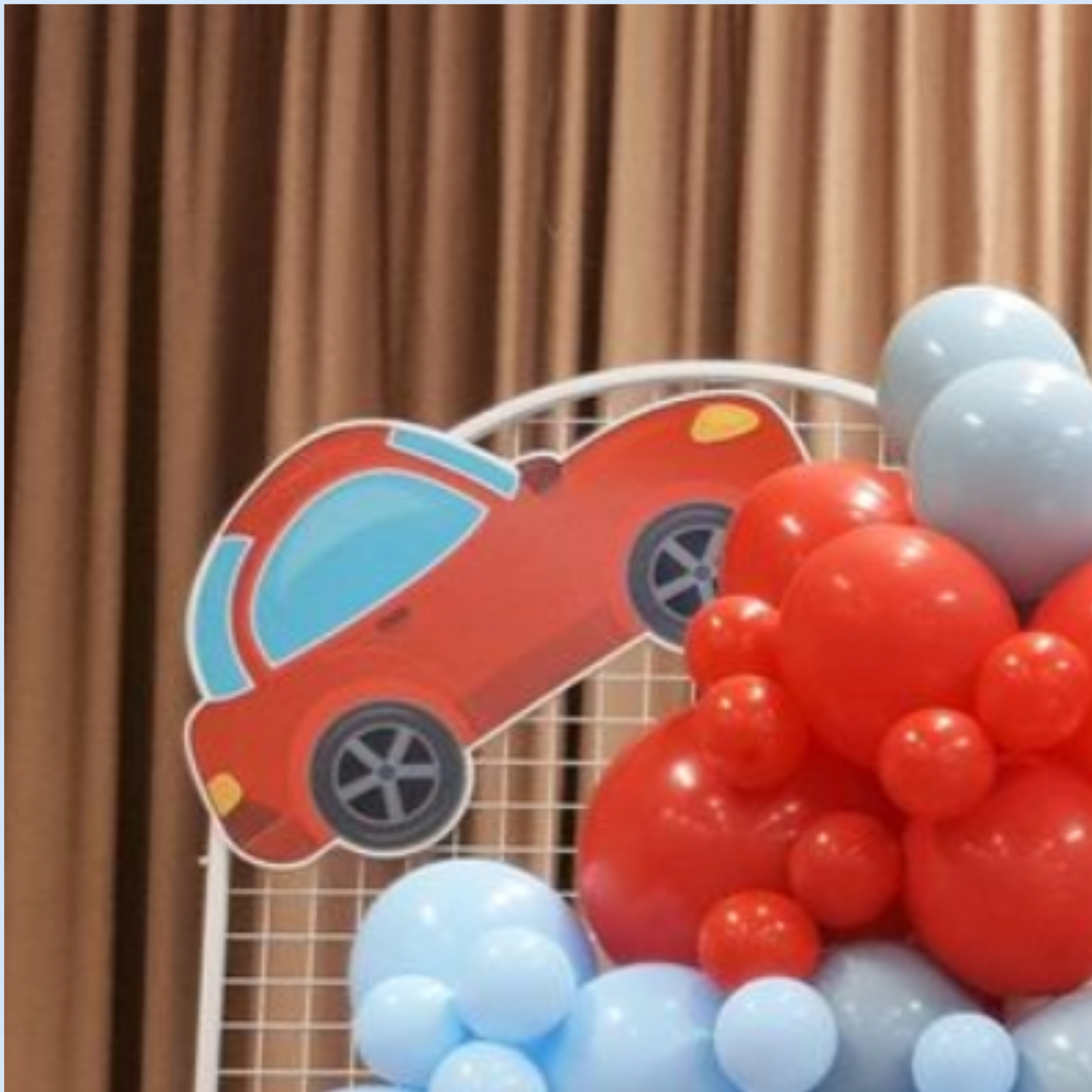 Vibrant red toy car cutout on a white wire grid, surrounded by clusters of red and blue balloons against a beige curtain backdrop. Ideal for children’s party decorations or toy store marketing. Lush and Lavish Events