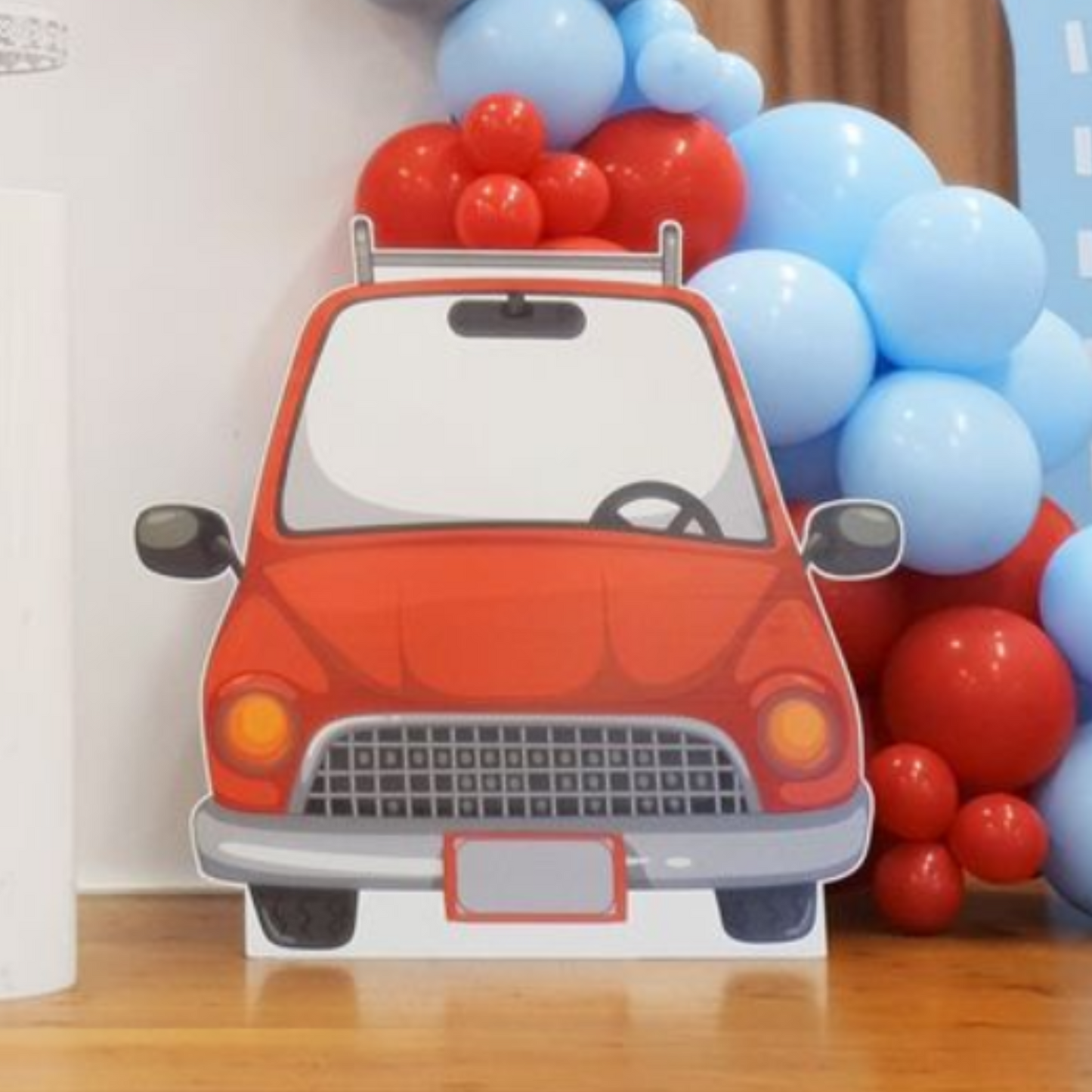 Red cartoon car cutout with big round headlights and mirrors in front of a blue and red balloon backdrop, perfect for a children’s party or automobile-themed celebration. Lush and lavish events