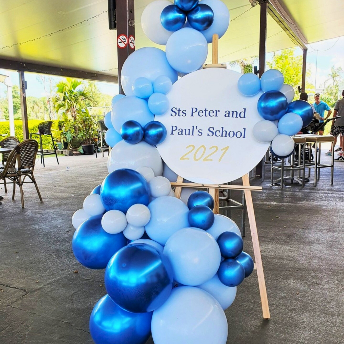 A celebratory display featuring a cluster of blue and white balloons arranged around a circular sign that reads ‘Sts Peter and Paul’s School 2021’. The arrangement is placed on an easel and is located in an outdoor setting with chairs and tables under a covered area. The festive decoration suggests a celebration or event related to the school, possibly a graduation or end-of-year ceremony for the year 2021. Prop hire, Prop hiring, Prop hire, event stylists Brisbane, brisbane event styling