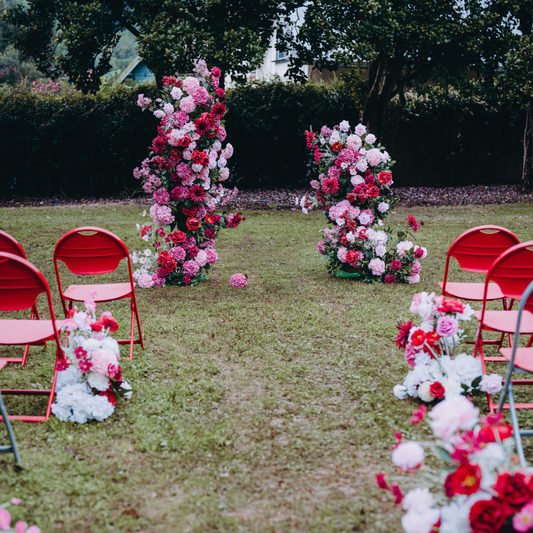 “An outdoor setting prepared for an event with rows of empty red chairs on either side of a grassy aisle. The aisle is lined with white and red flowers leading to an arch made of similar flowers at the end, creating a picturesque scene for a ceremony.