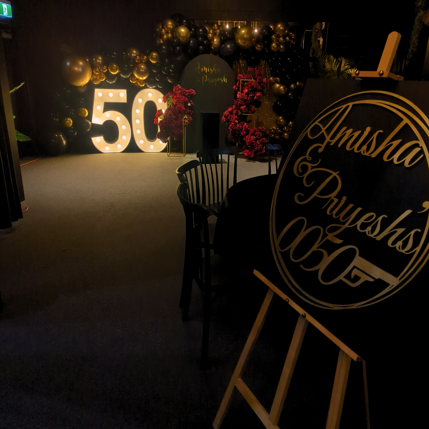An indoor celebration venue with dim lighting and a festive atmosphere. In the background, large golden balloons form the number ‘50’, surrounded by smaller balloons and lights. 