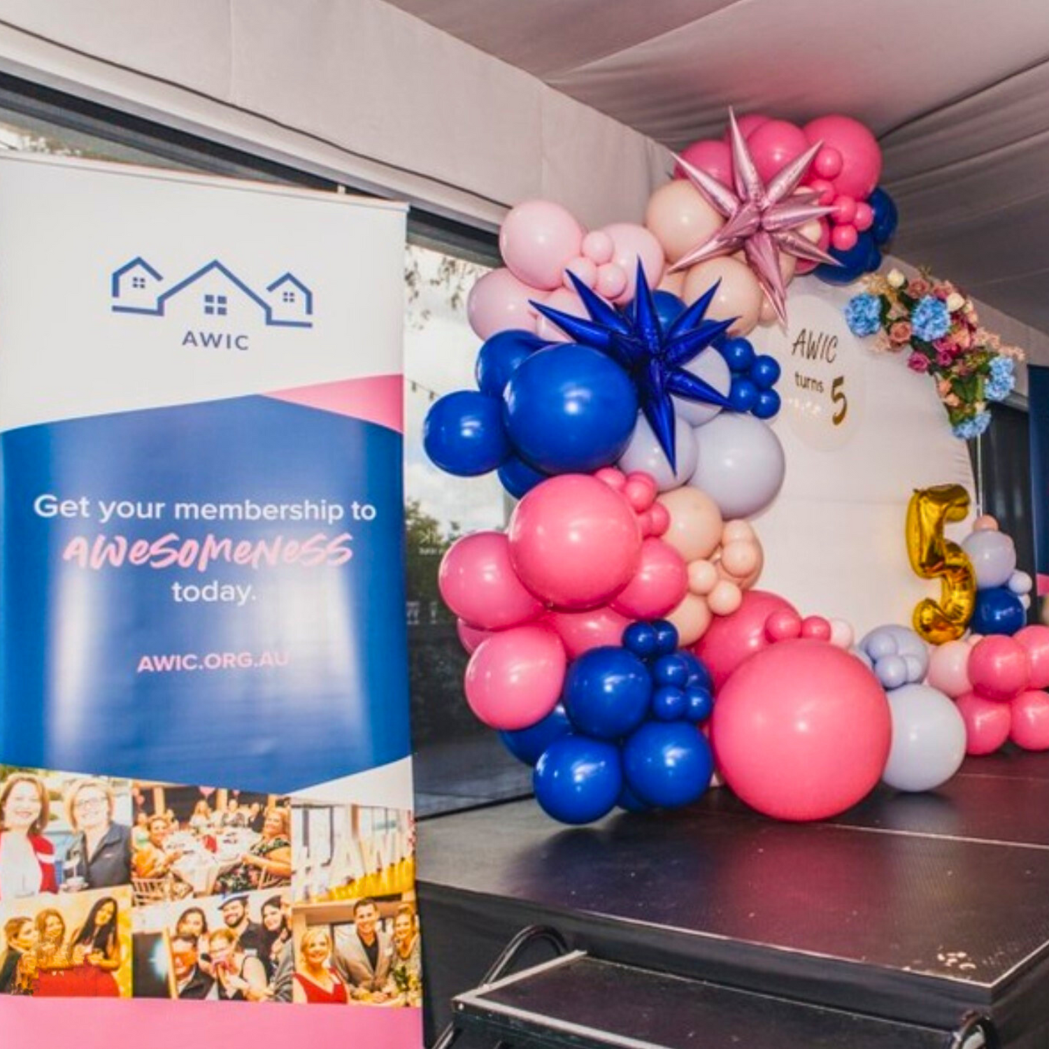 A vibrant event setup featuring a banner with ‘AWIC’ and ‘Get your membership to AWESOMENESS today. AWIC.ORG.AU’ alongside event images. The banner is flanked by colorful balloon arrangements in pink, blue, and purple hues with a large golden ‘5’ balloon.