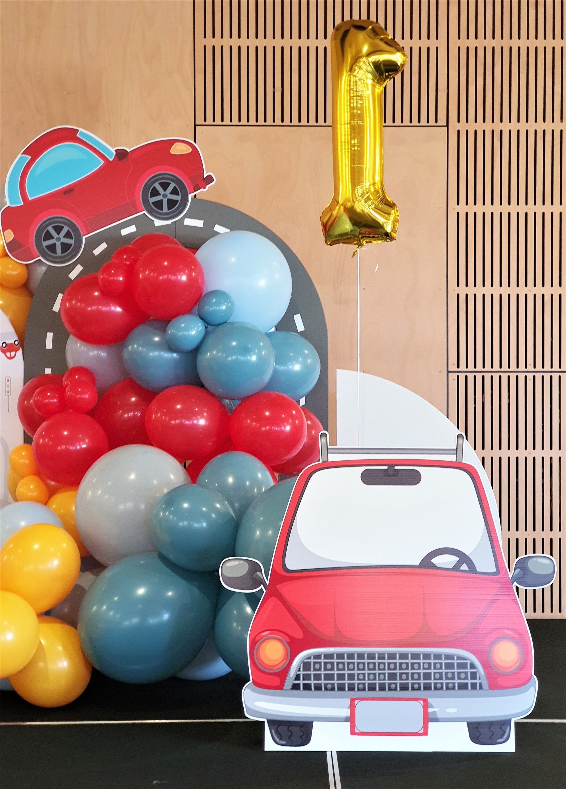 A colorful car-themed birthday party setup with red, blue, and yellow balloons. There's a large cutout of a red car, a golden number "1" balloon, and a smaller car cutout in the background. The setting appears to be indoors. Prop hire, Prop hiring, Prop hire, event stylists Brisbane, brisbane event styling, brisbane event styling, event stylist Brisbane, Brisbane Wedding, Gold Coast Wedding, Floral Styling, Lush & Lavish Events
