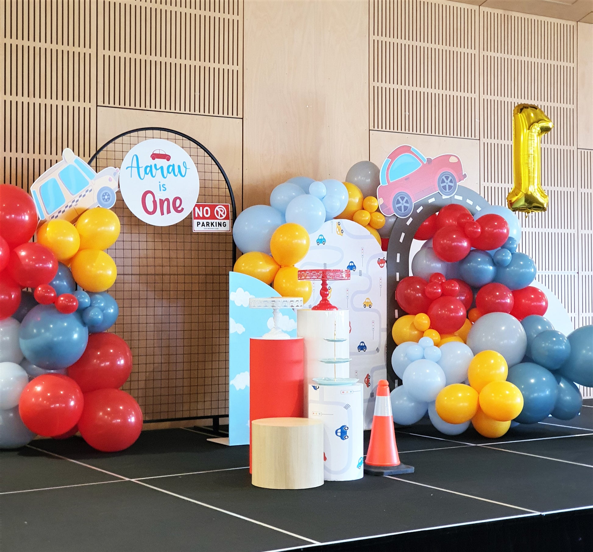 A first birthday stage decorated with colorful car-themed balloons, a sign reading "Aarav is One," and a large gold number one balloon. Props include traffic cones and cylindrical stands. Prop hire, Prop hiring, Prop hire, event stylists Brisbane, brisbane event styling, brisbane event styling, event stylist Brisbane, Brisbane Wedding, Gold Coast Wedding, Floral Styling, Lush & Lavish Events