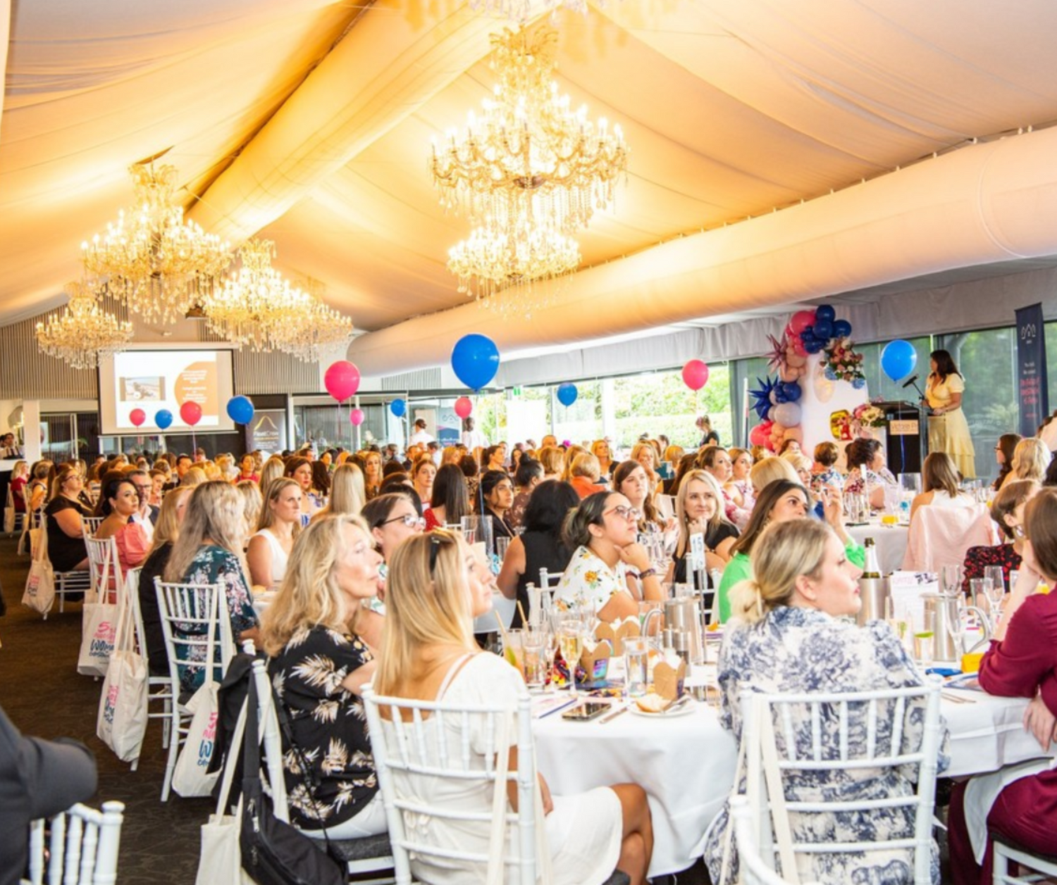 Event setting with white-clothed tables, blue and pink balloon centerpieces, chandeliers, and a presentation screen. Prop hire, Prop hiring, Prop hire, event stylists Brisbane, brisbane event styling, brisbane event styling, event stylist Brisbane, Brisbane Wedding, Gold Coast Wedding, Floral Styling, Lush & Lavish Events, Brisbane Corporate Styling, Gold Coast Corporate Styling, Brisbane Brand Activation